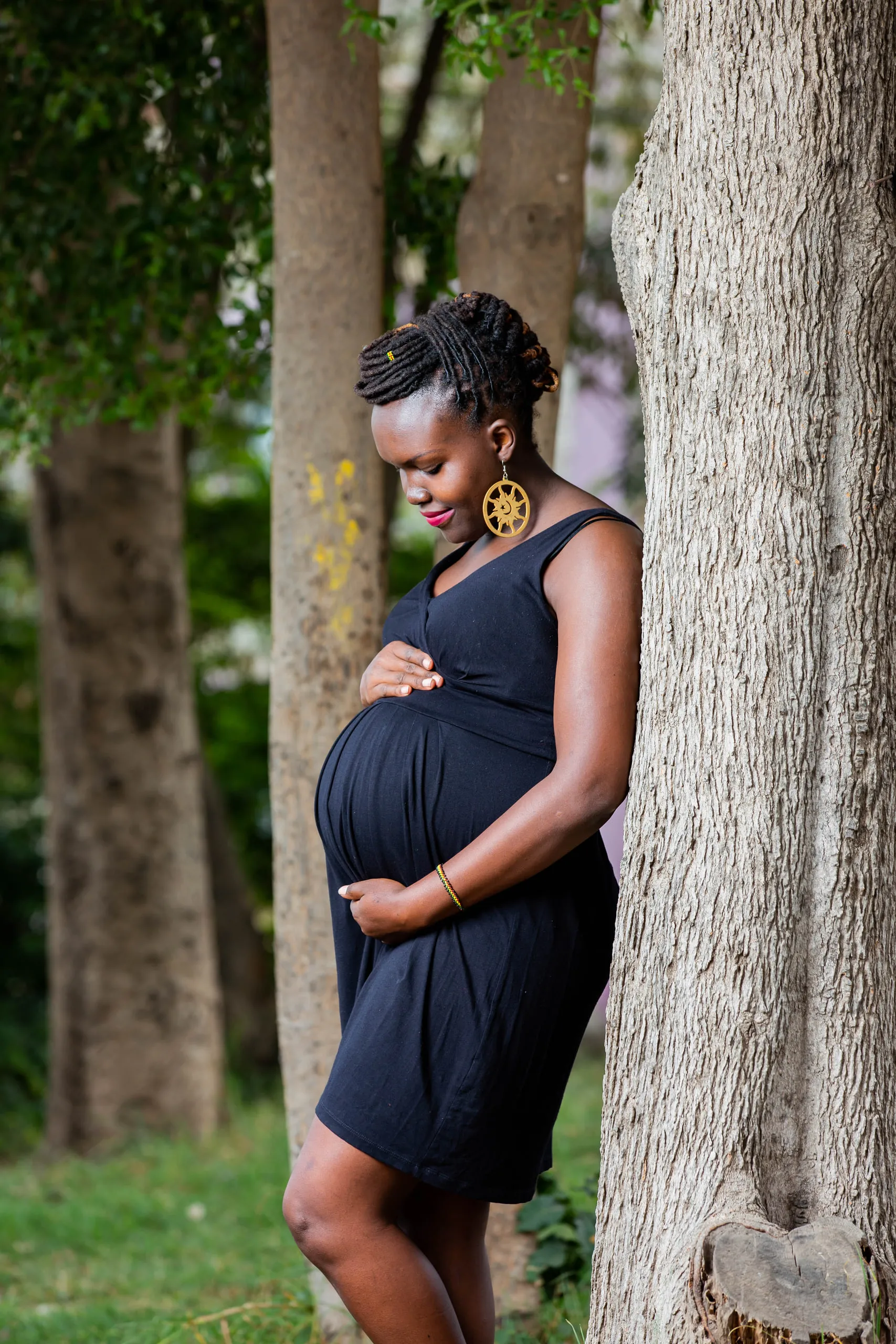 Pregnant woman in a black dress leaning against a tree trunk with her hands on her belly