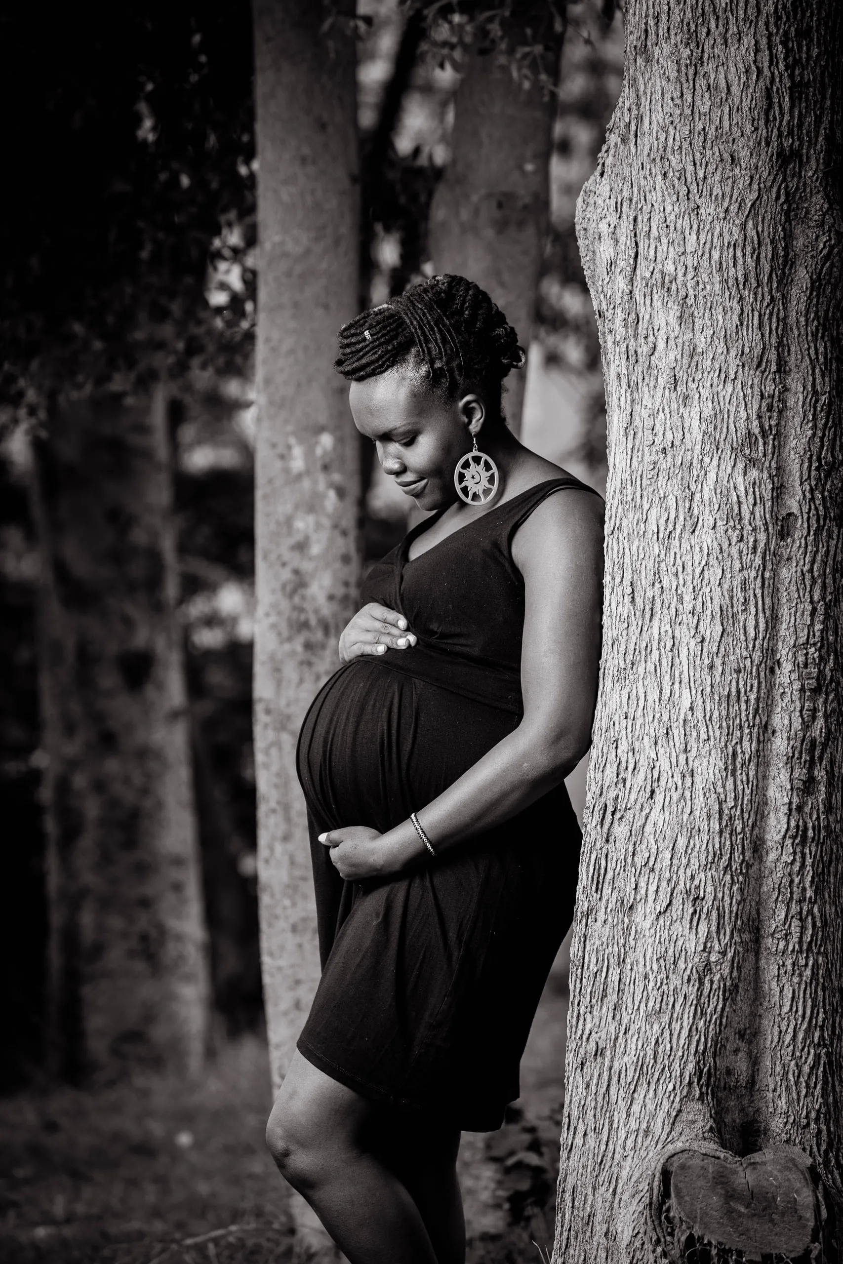 Pregnant woman in a black dress leaning against a tree, hands on her belly, in black and white
