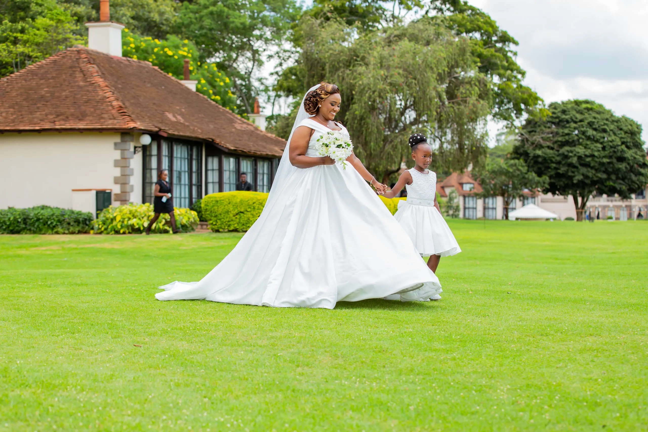 Bride in a full gown crossing a wide lawn with a flower girl