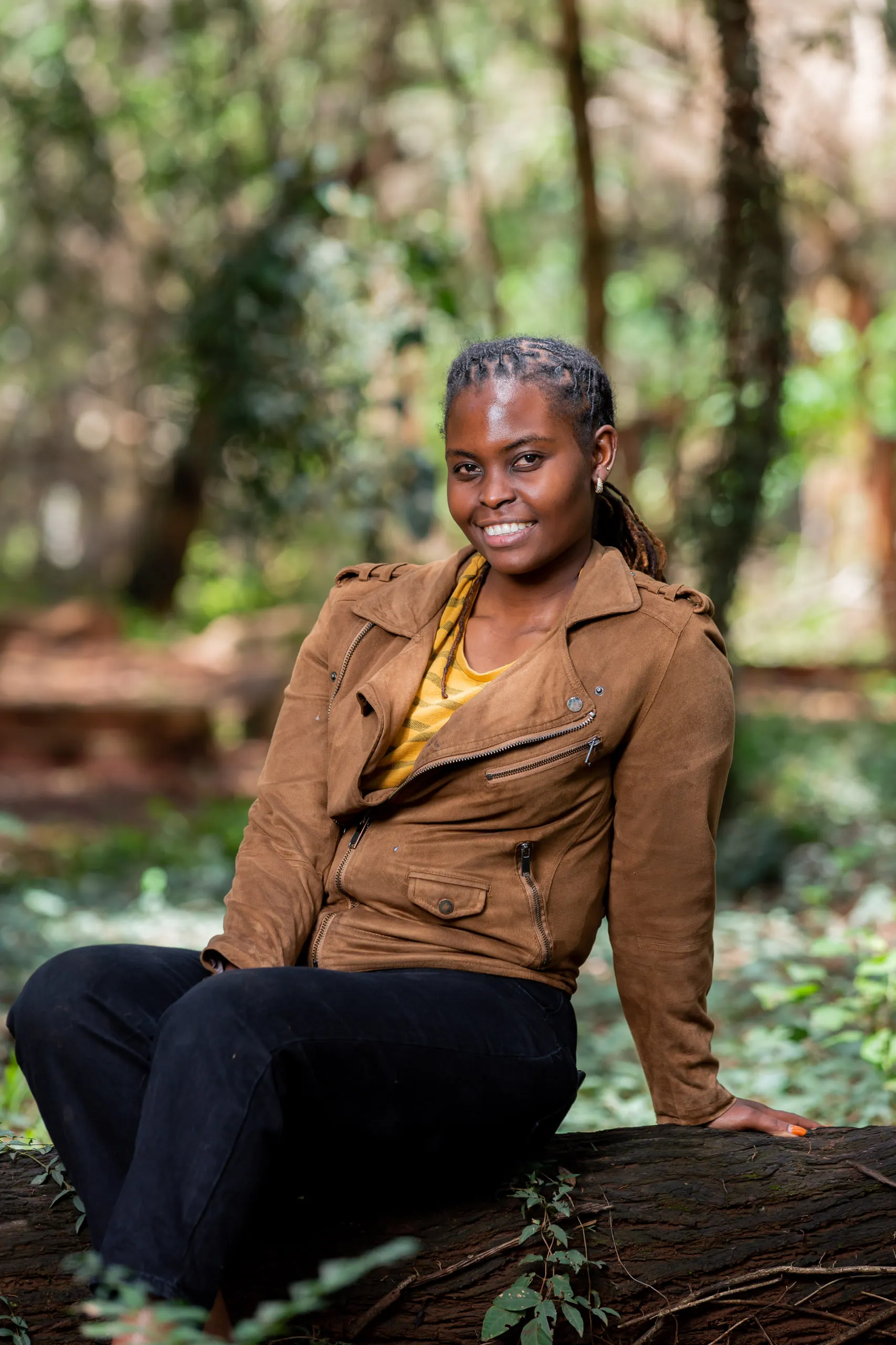 Woman in a tan suede jacket smiling as she sits on a fallen log in the forest