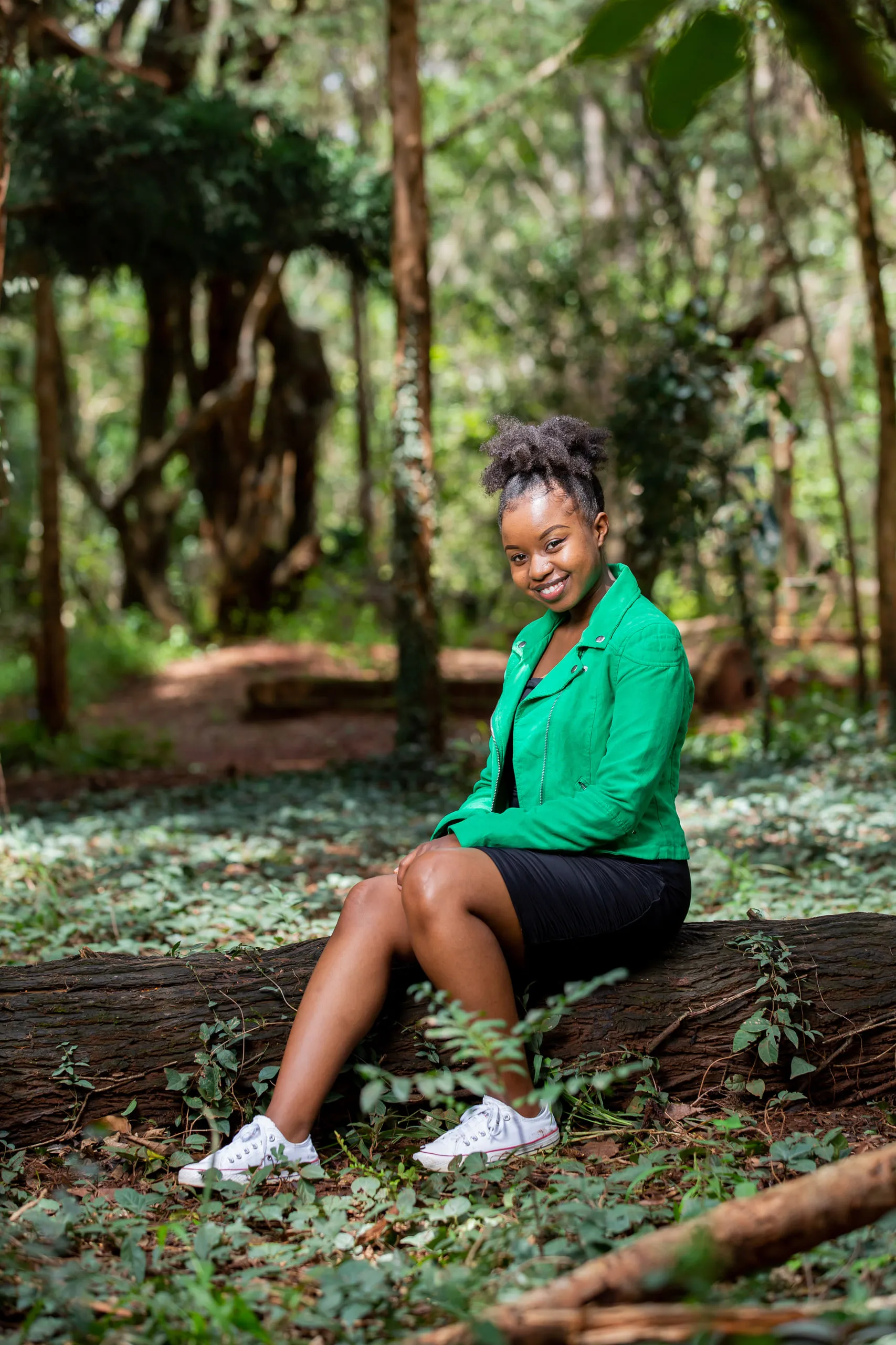 Woman in a green jacket and white trainers sitting on a log among ivy, smiling at the camera
