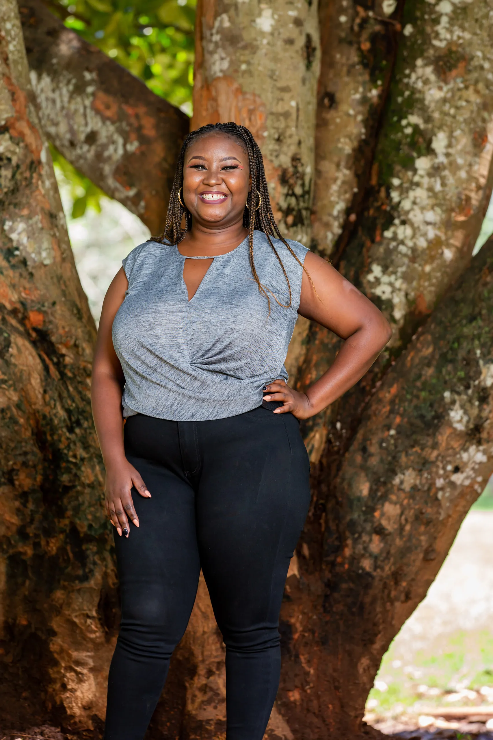 Woman in a grey top and black trousers with a hand on her hip, standing in front of a large tree trunk