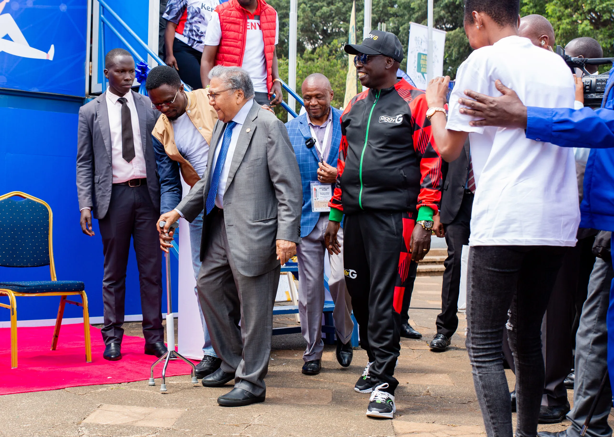 Elderly man with a walking stick crossing a red carpet among a crowd of officials