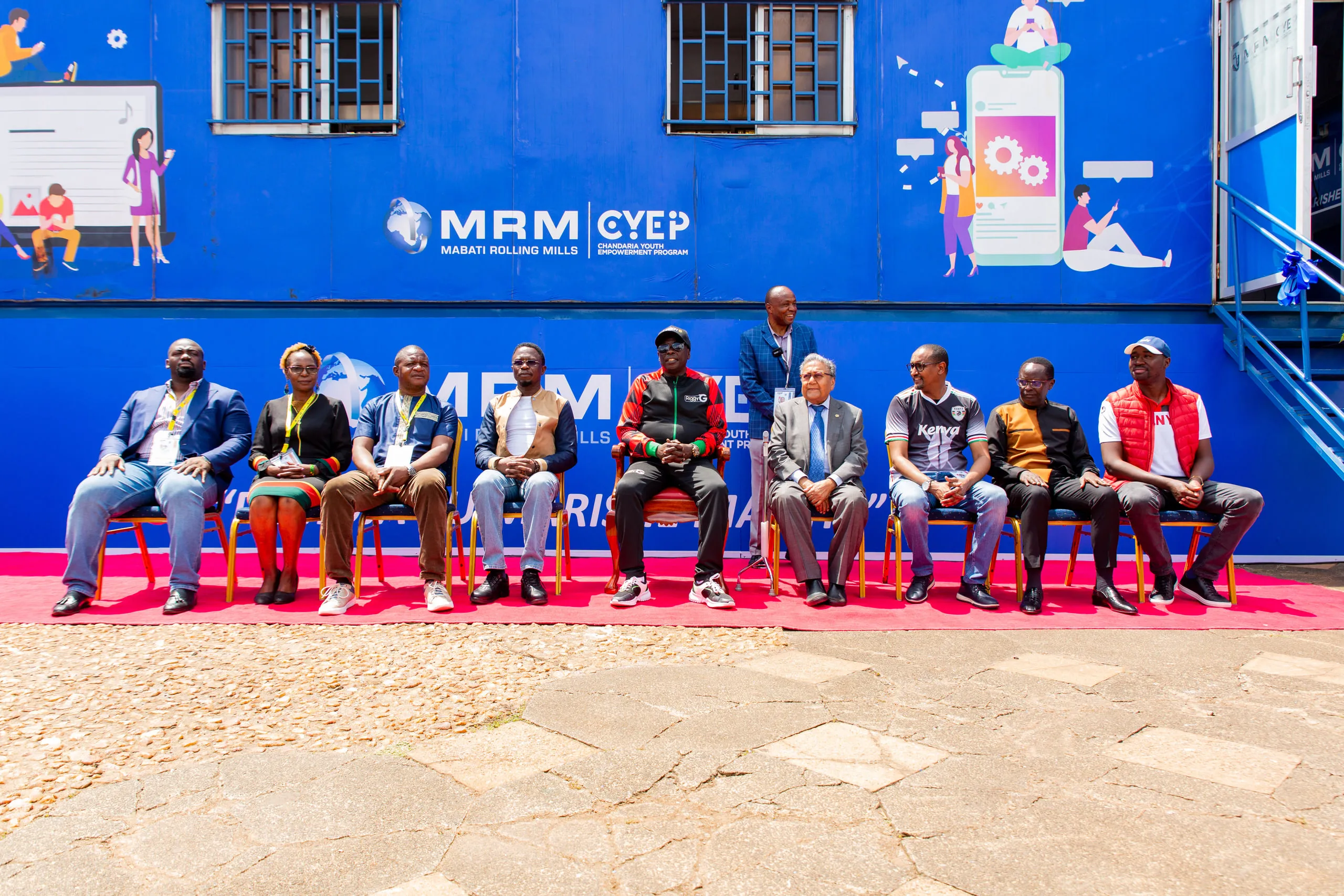Dignitaries seated in a row on a red carpet in front of a blue branded container