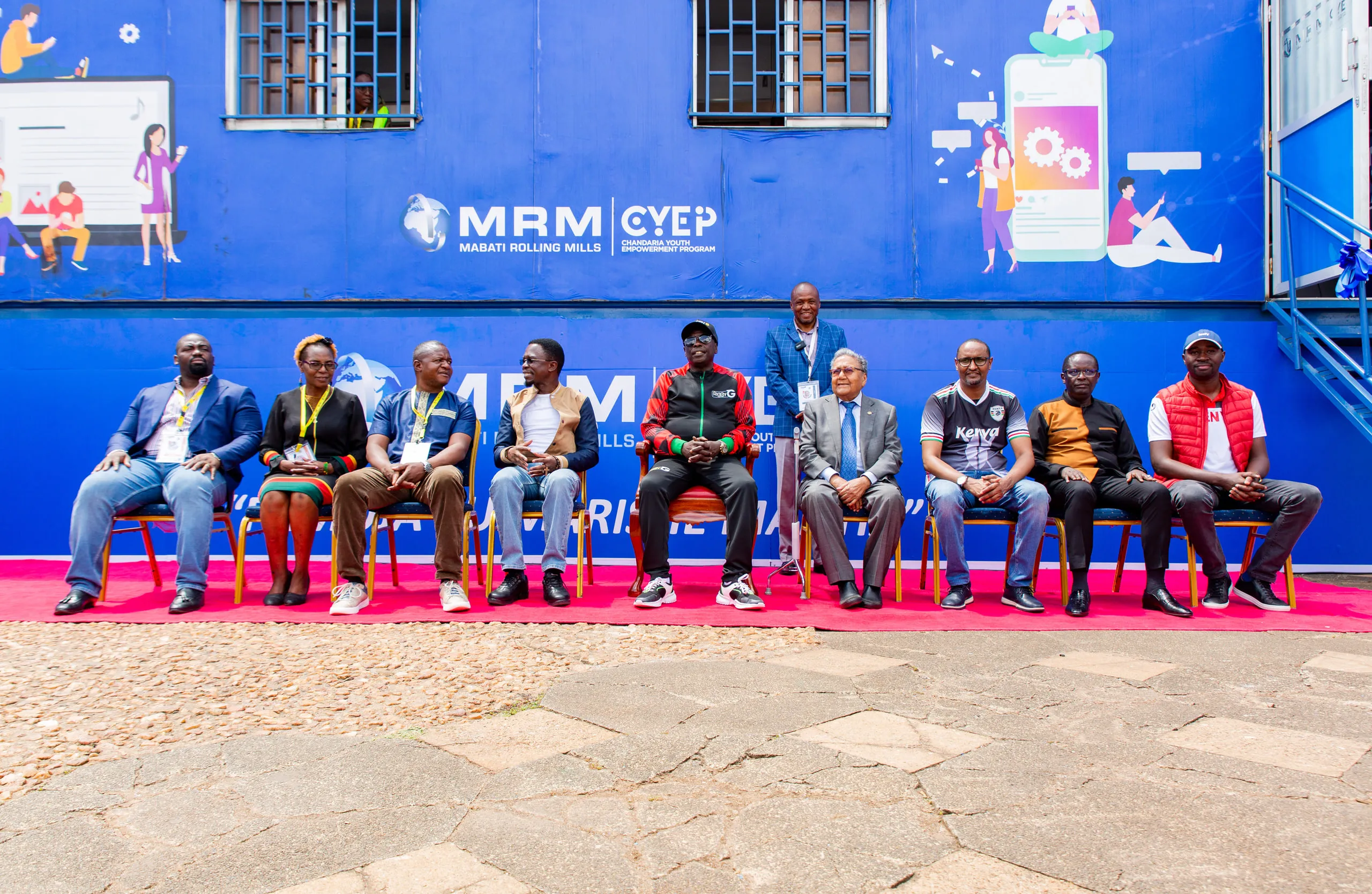 Row of dignitaries seated on a red carpet with one man standing behind them