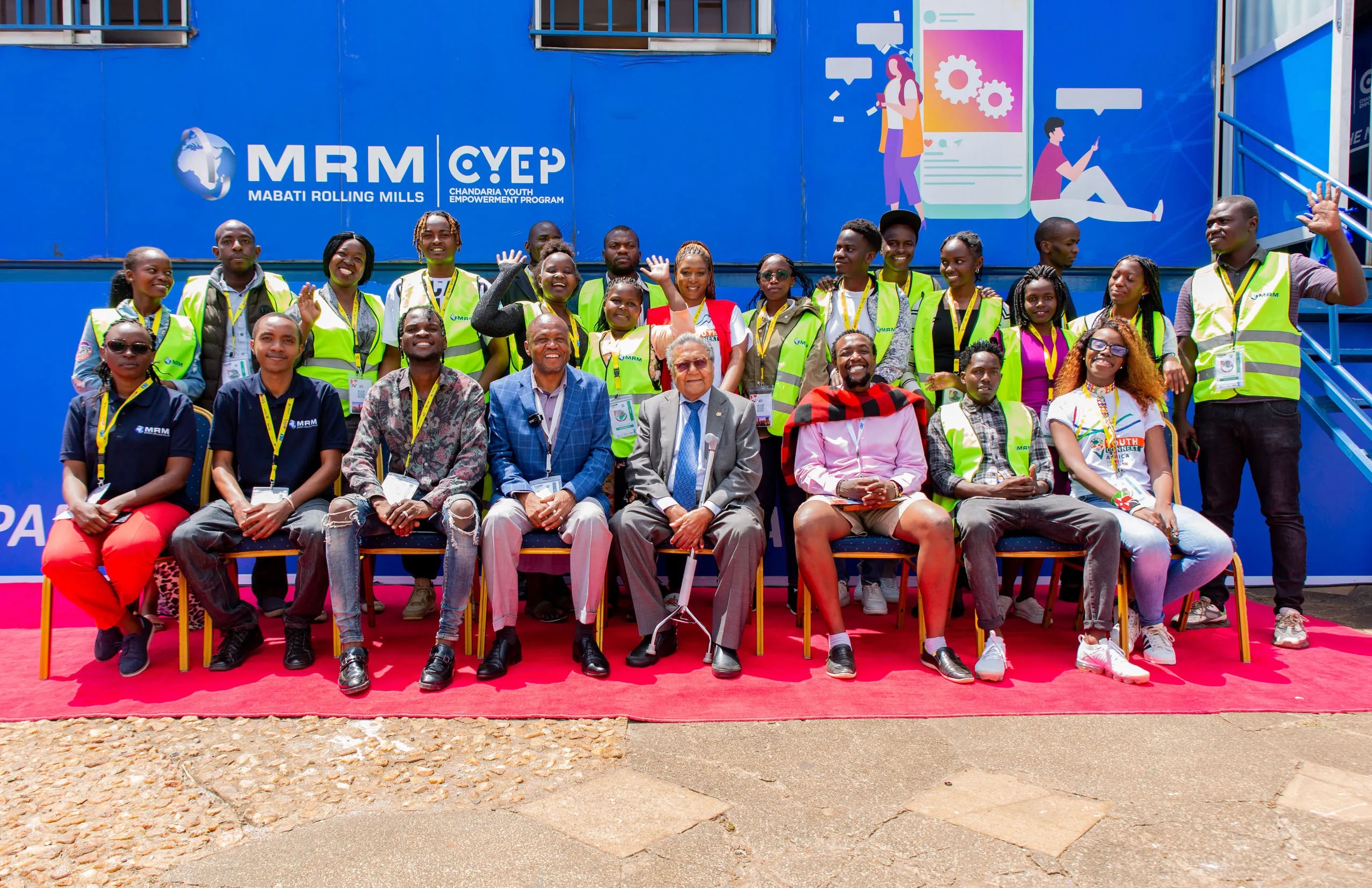 Seated dignitaries on a red carpet with volunteers in green vests standing behind them