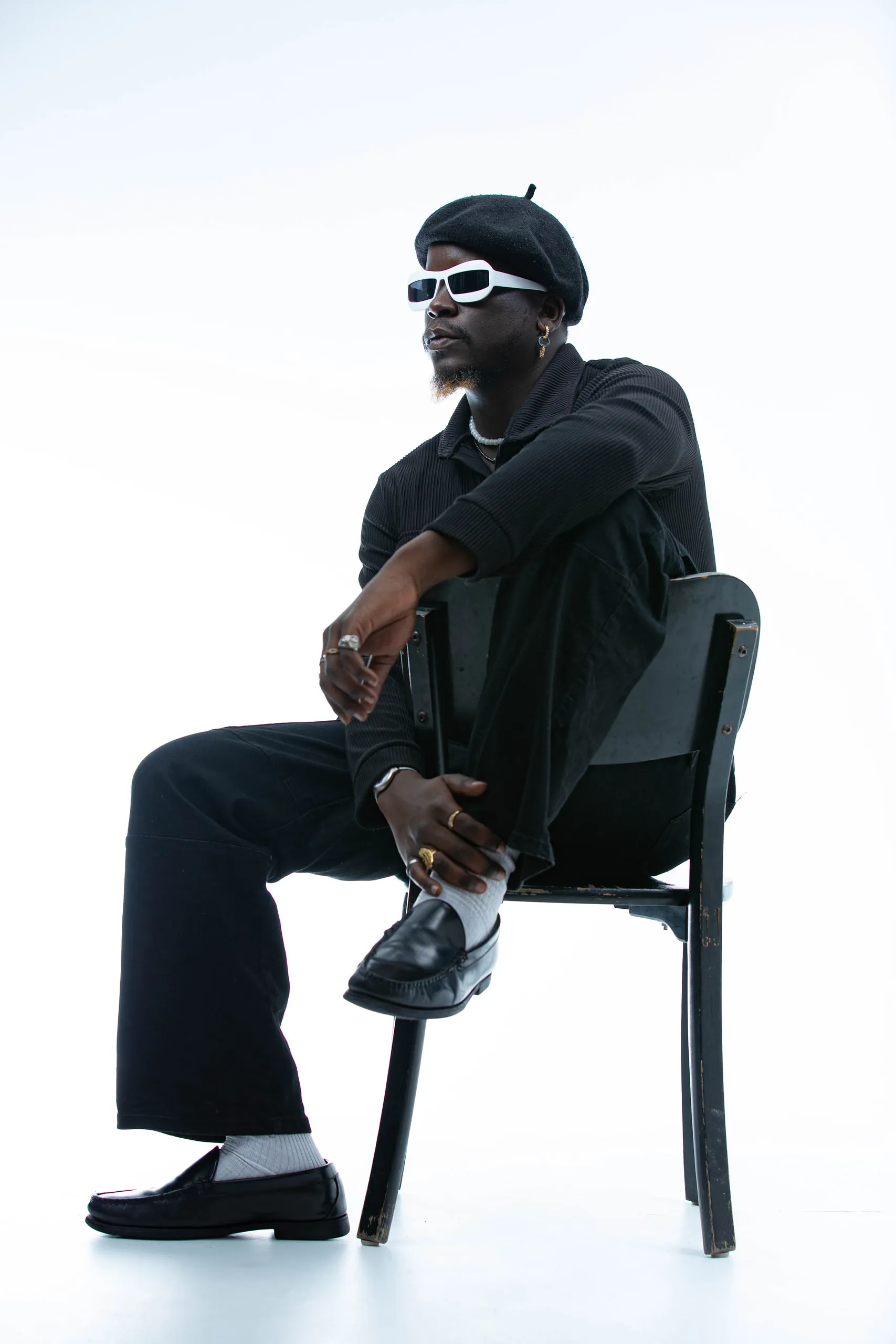 Man in a black beret and white sunglasses sitting on a chair with one foot on the seat, on white