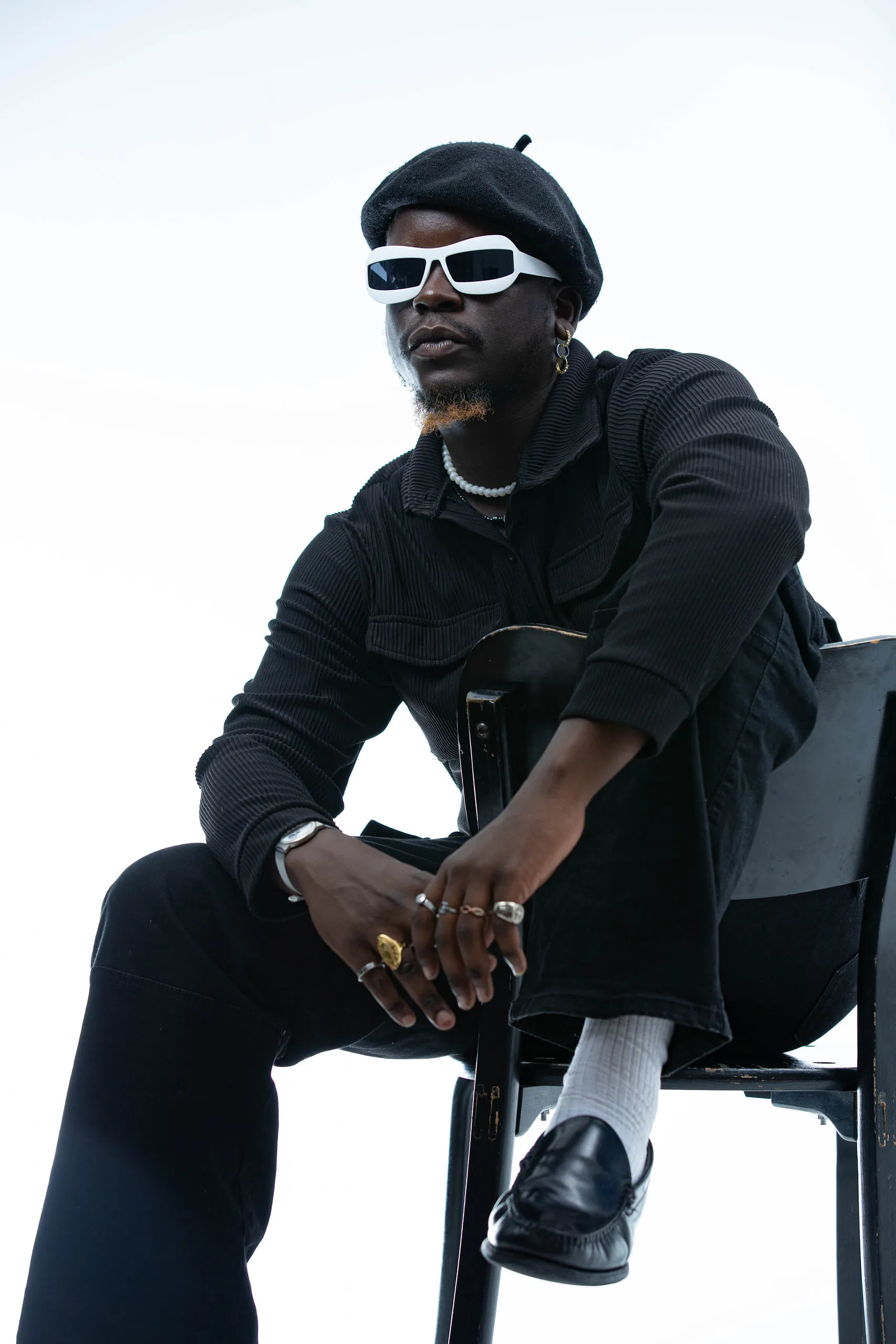 Man in a black beret and white sunglasses seen from below, hands clasped over his knee