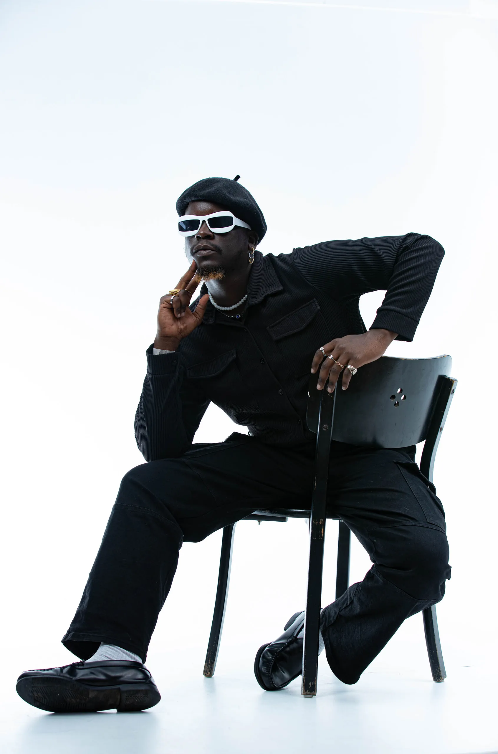 Man in a beret and white sunglasses touching his chin, leaning over the back of a chair