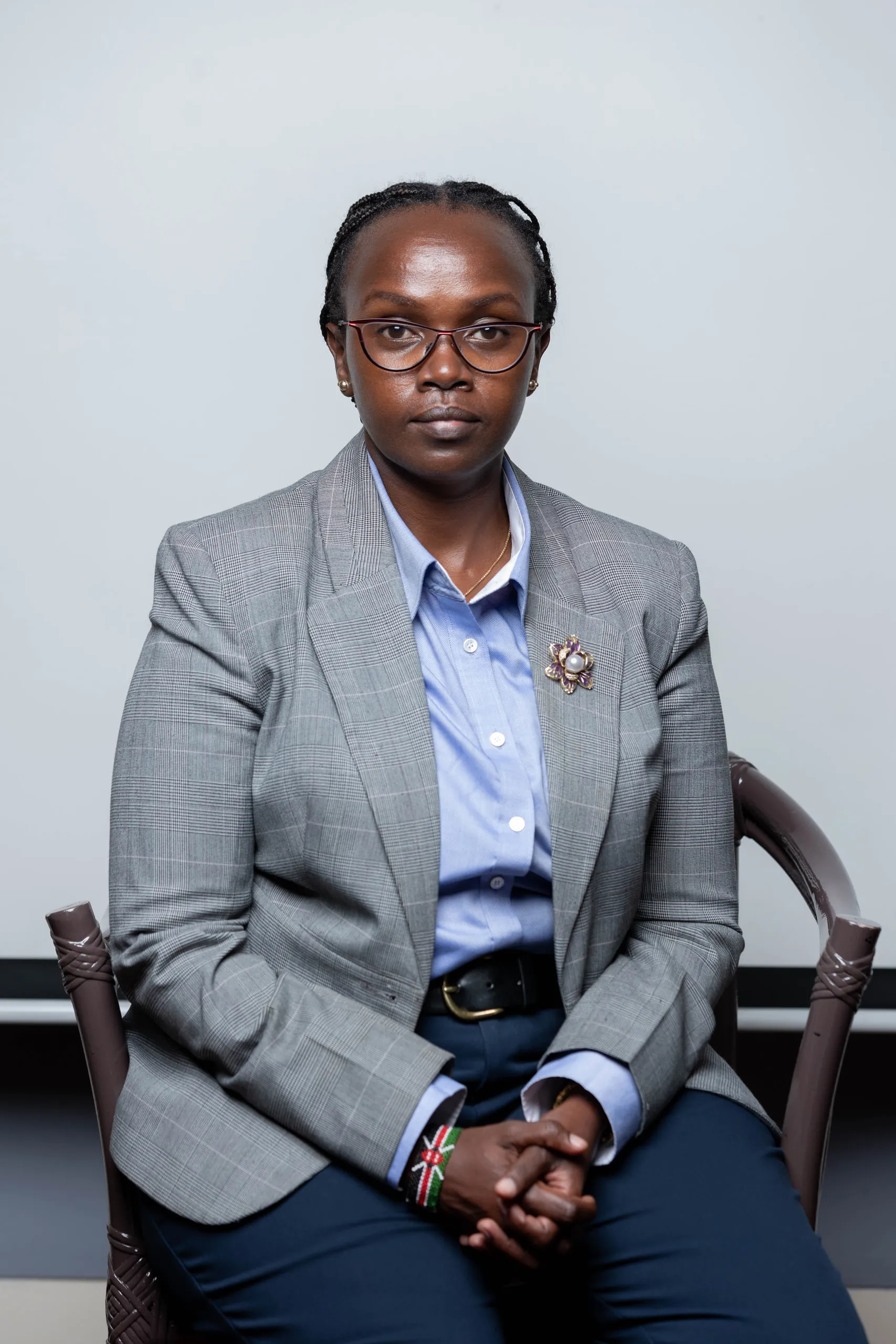 Woman in a grey blazer and glasses sitting with her hands clasped against a pale wall