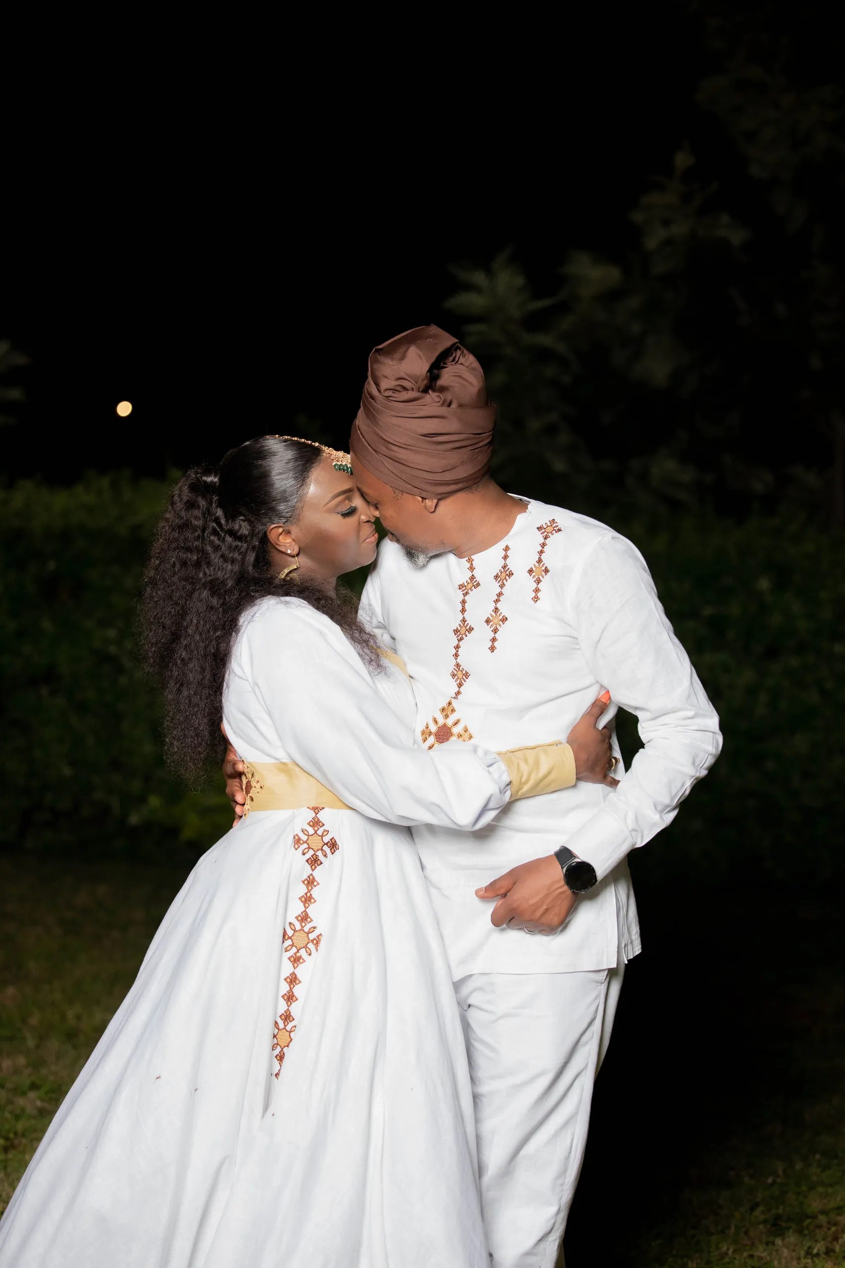 Bride kissing the groom's cheek at night, he in a headwrap