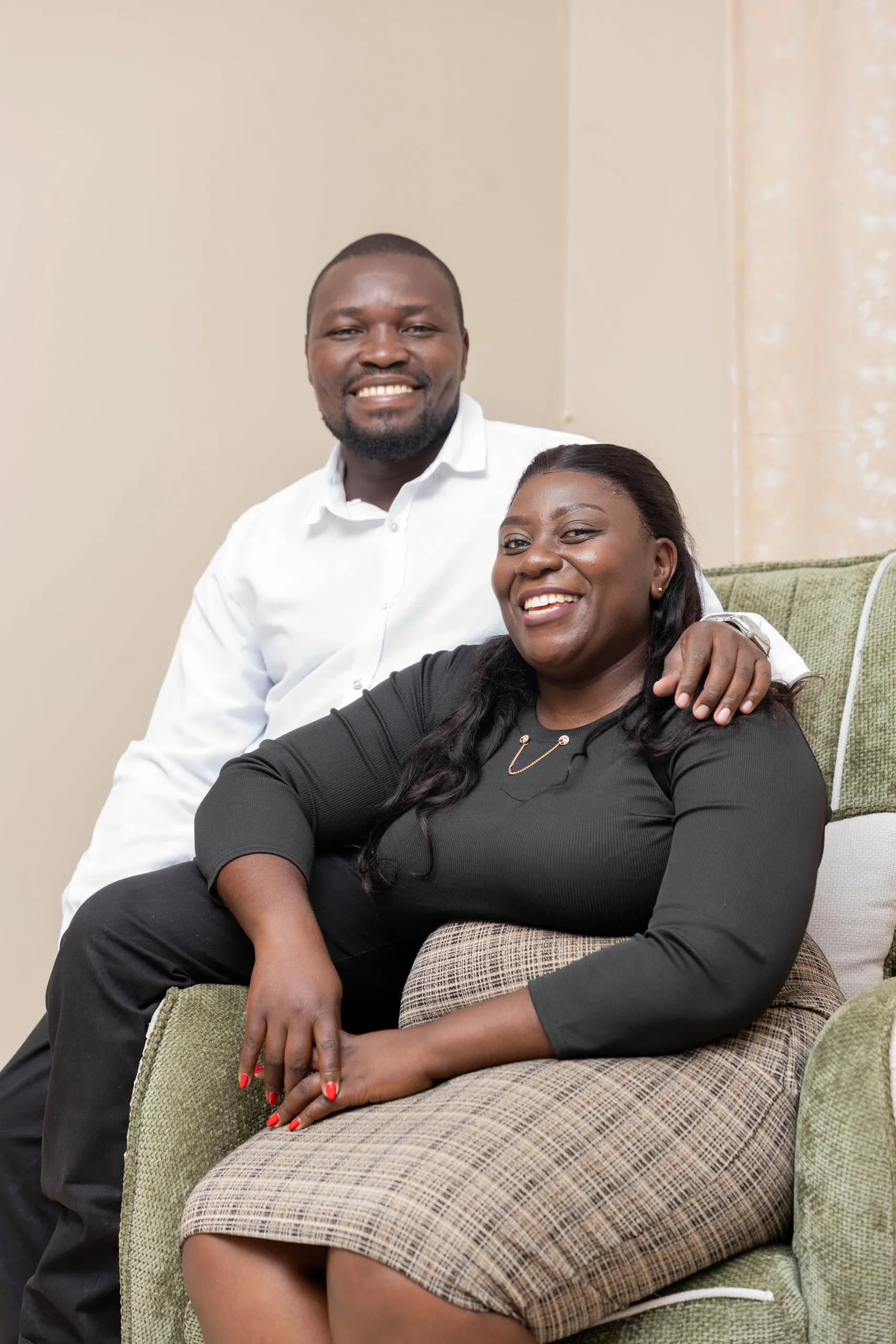 Couple smiling on a green armchair, he in a white shirt standing behind her, she in a grey dress