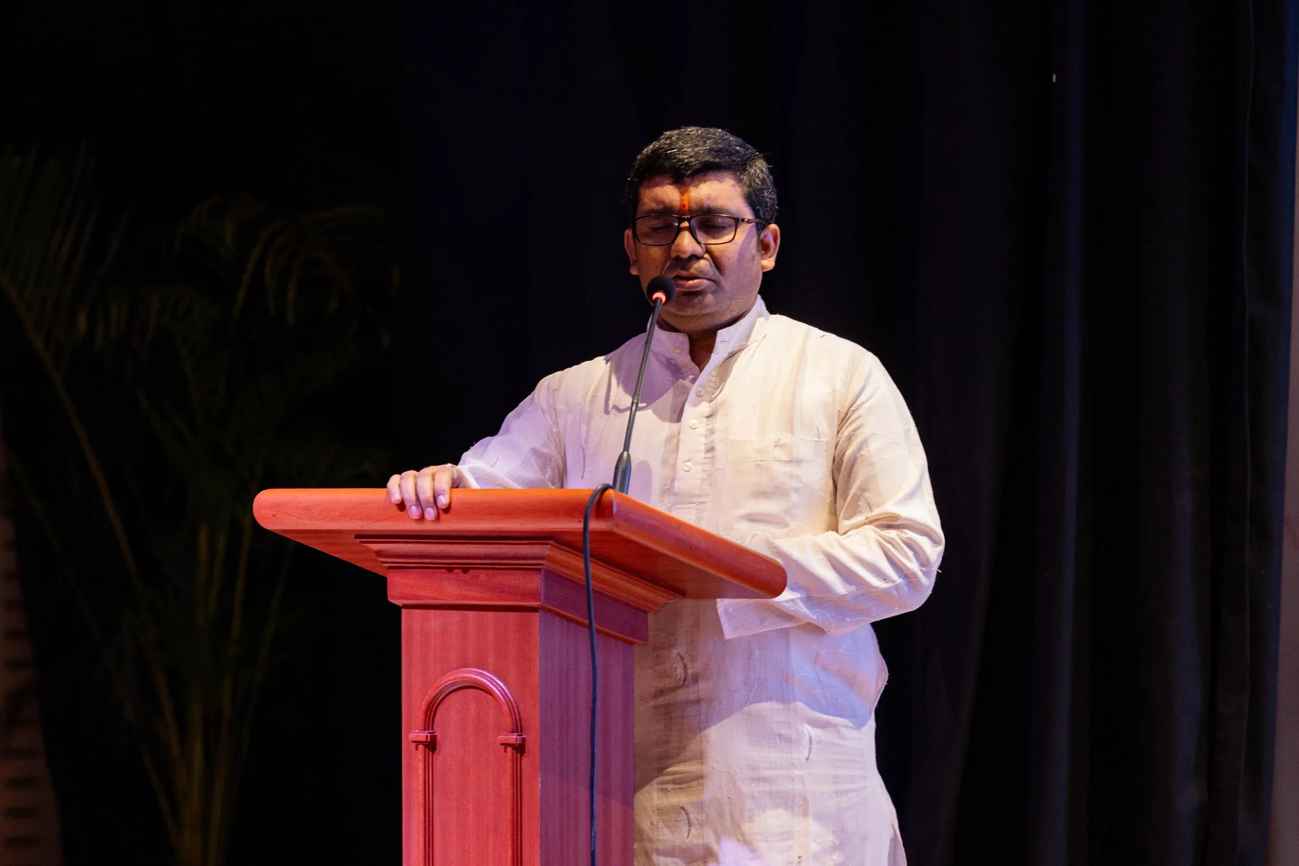 Man in a white kurta speaking at a wooden lectern against a dark curtain
