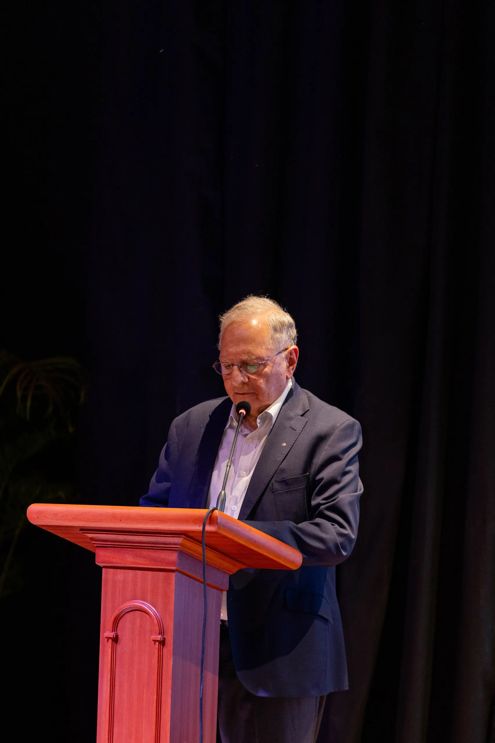 Older man in a navy blazer reading at a wooden lectern against a dark curtain