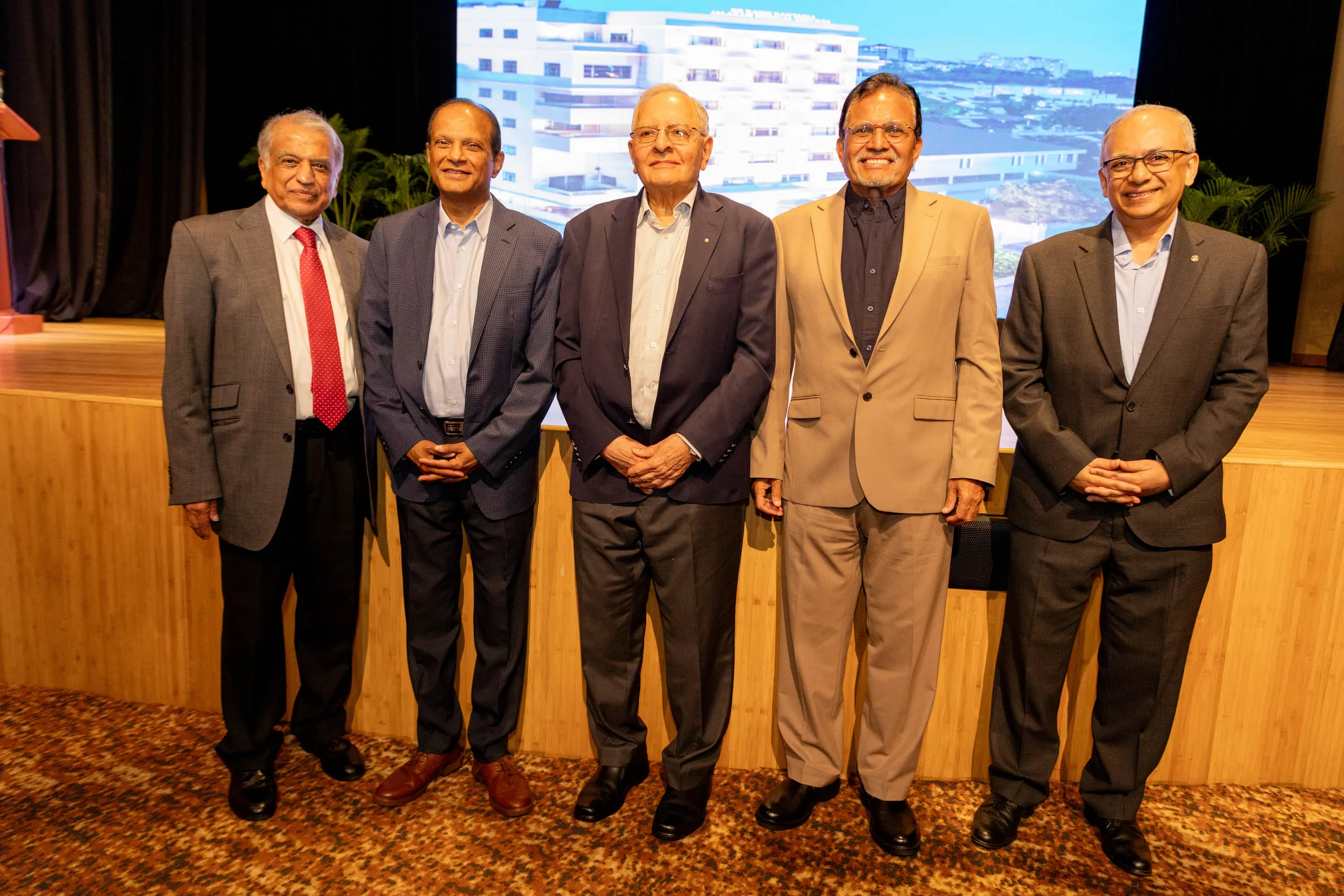 Five men in jackets standing in a row in front of a stage and projected screen