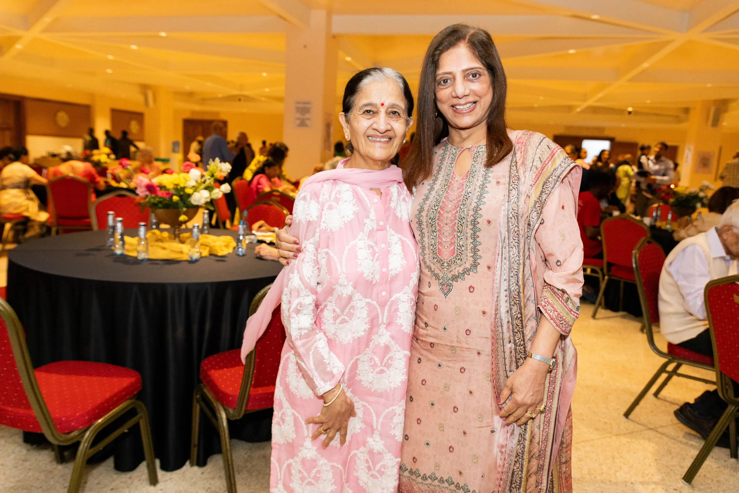 Two women in pink traditional outfits embracing in front of a dining hall with flowers