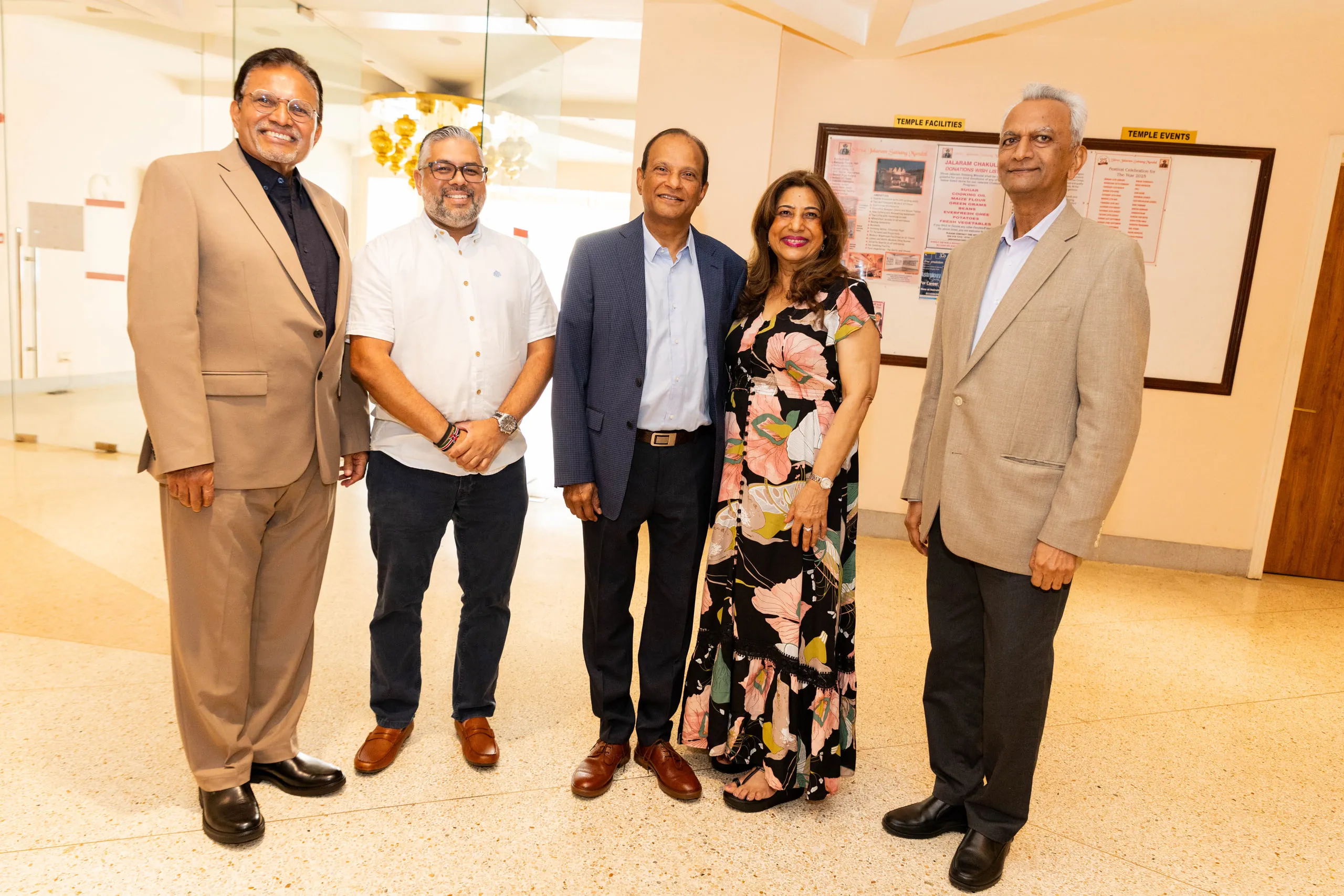 Five adults in smart clothes posing together in a corridor with a notice board