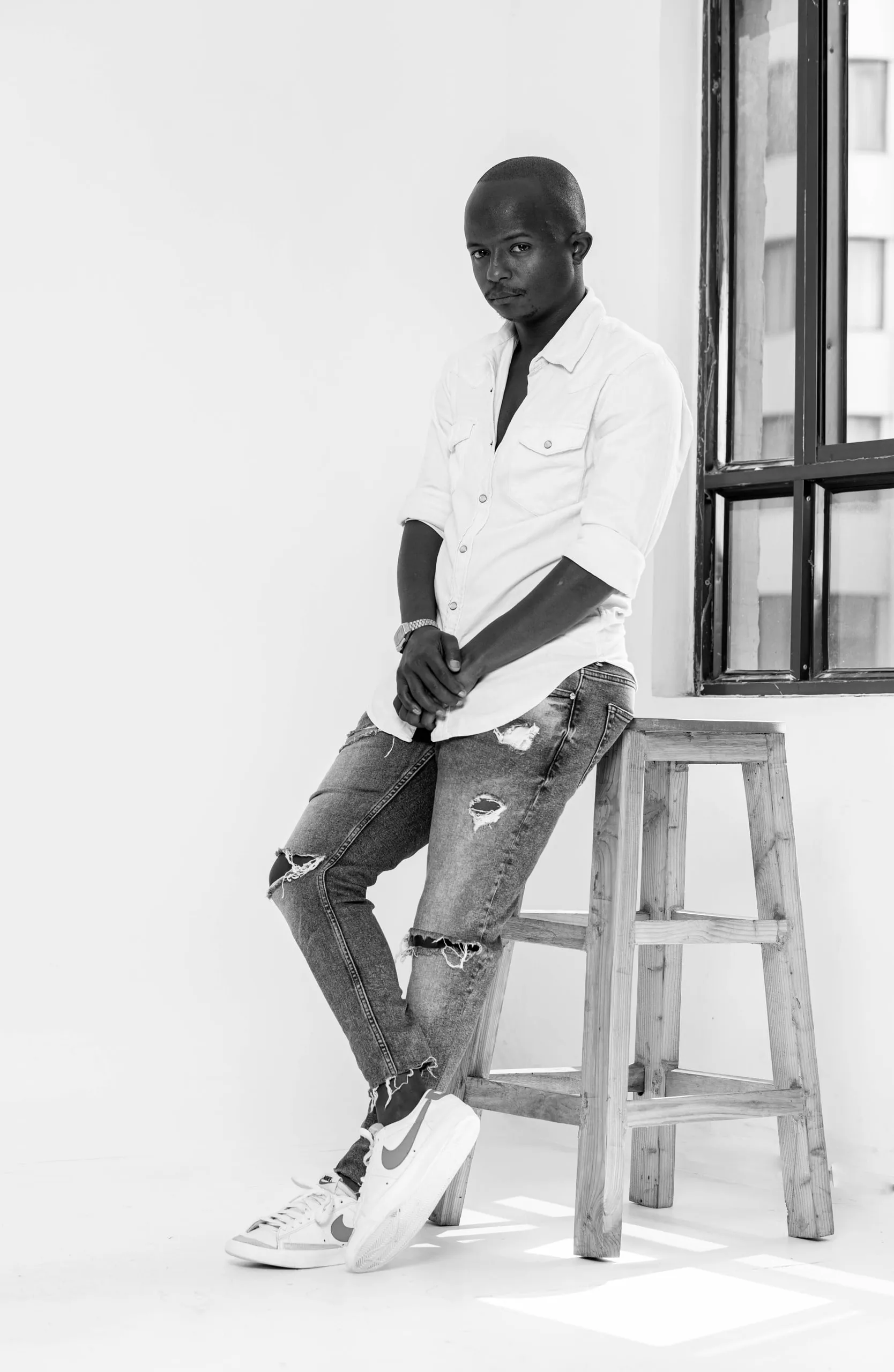 Man in a white shirt and ripped jeans leaning on a stool by a window, in black and white