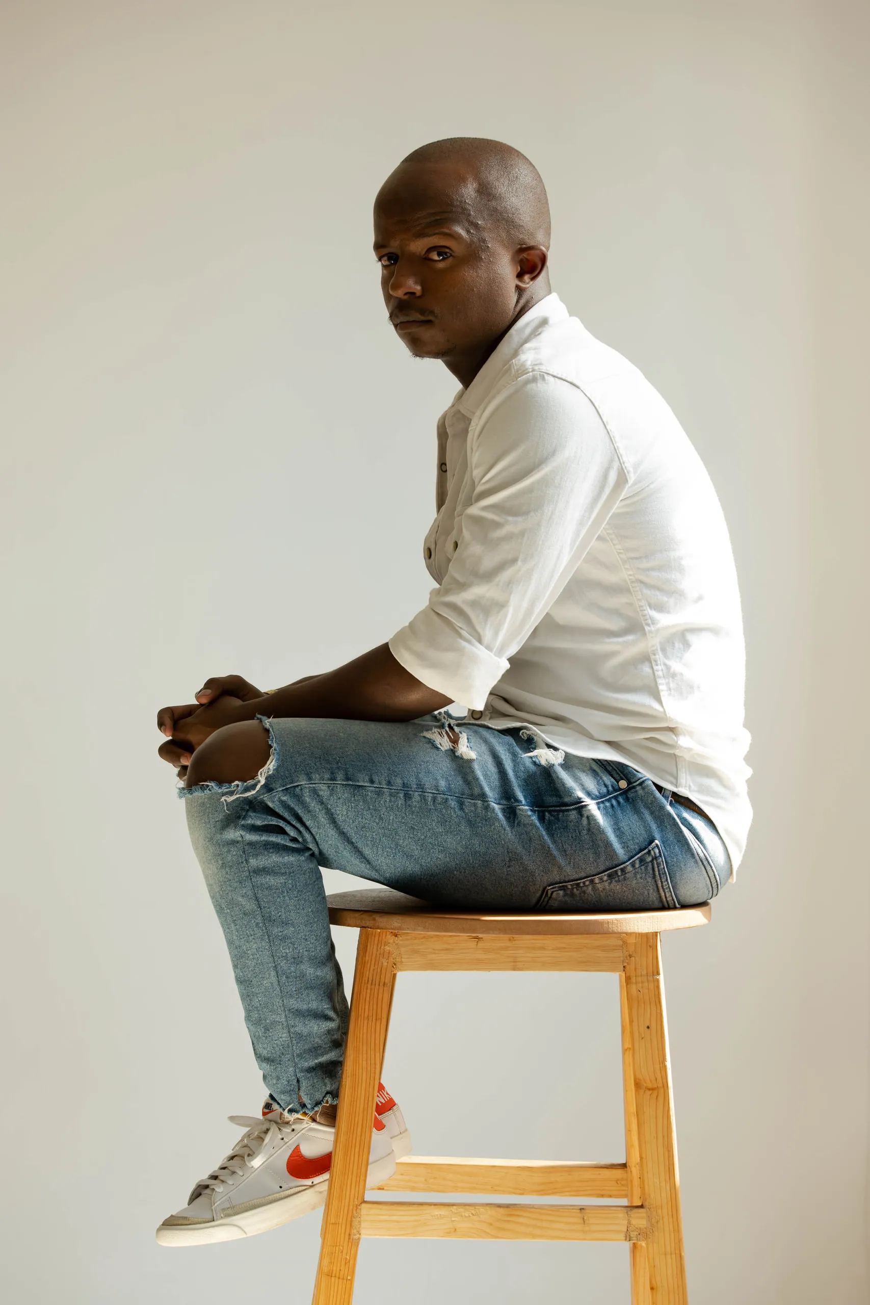 Man in a white shirt and ripped jeans sitting on a wooden stool, looking back at the camera