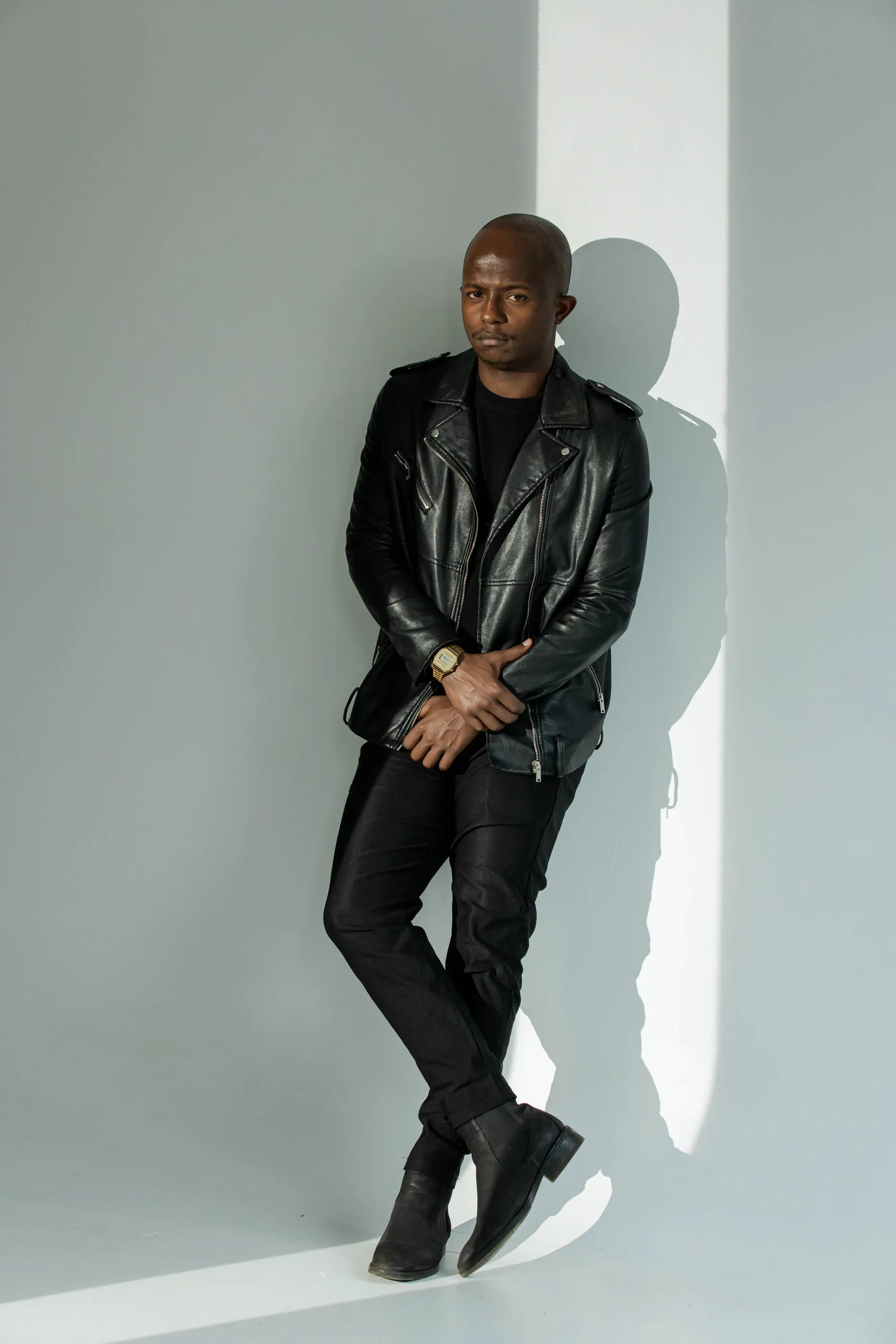 Man in a black leather jacket leaning on a pale wall beside a bar of sunlight