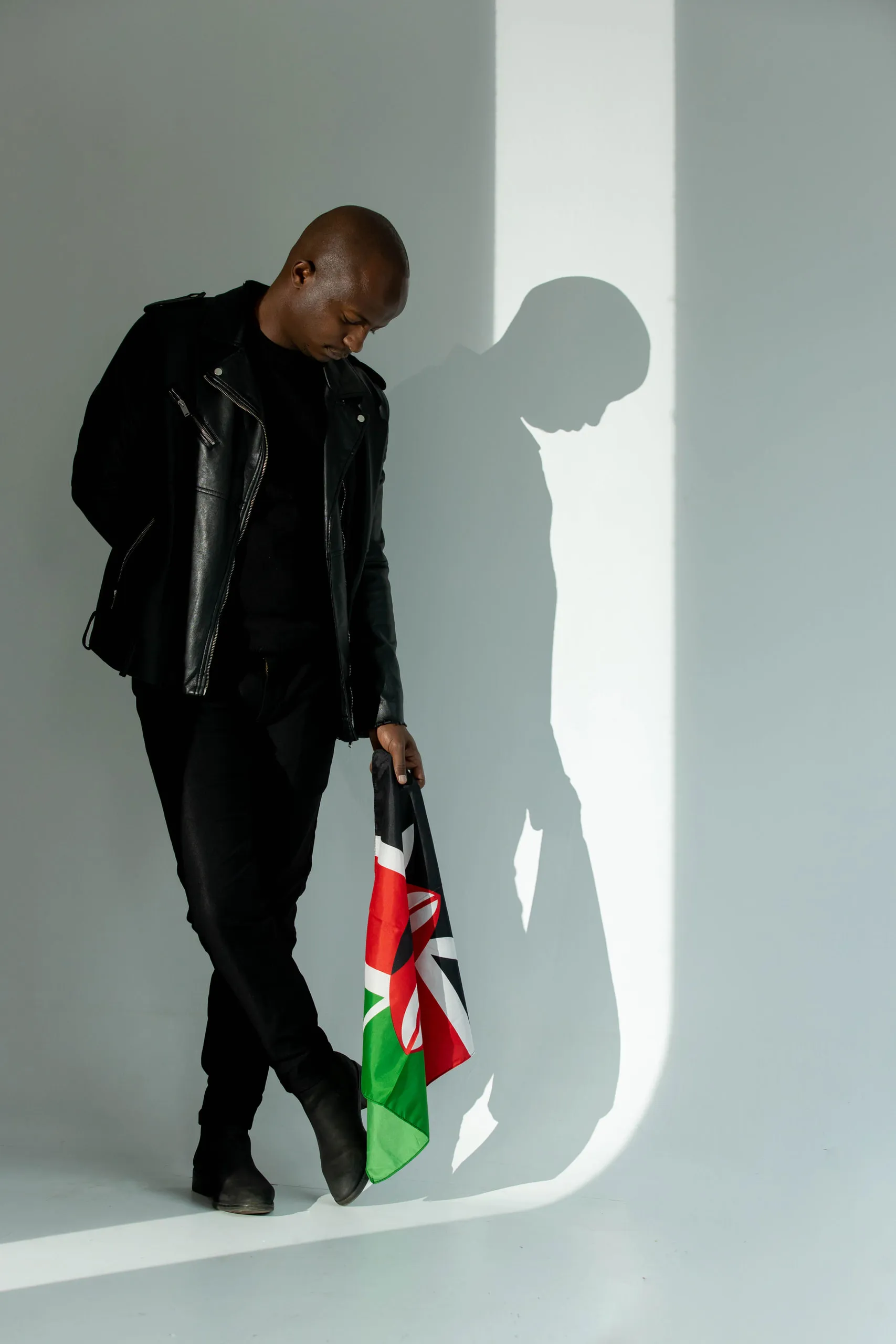 Man in a leather jacket looking down and holding a Kenyan flag, with his shadow on the wall