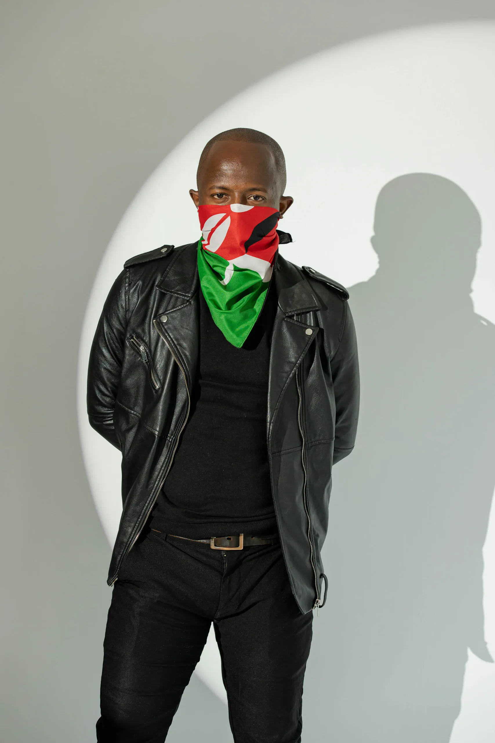 Man in a black leather jacket with a Kenyan flag bandana over his face, standing in a pool of light