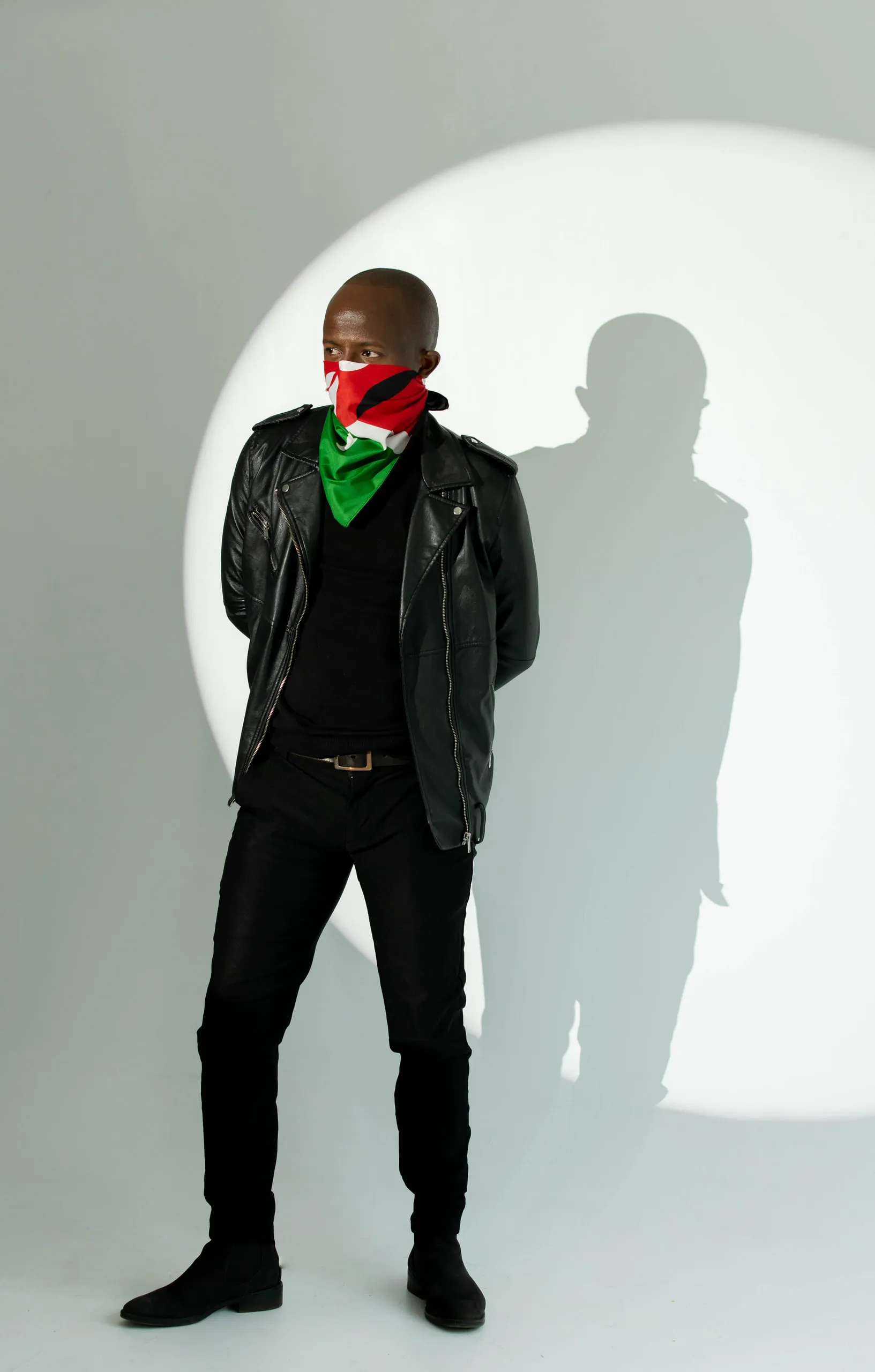Man in a leather jacket and flag bandana looking sideways in front of a round spotlight and his shadow