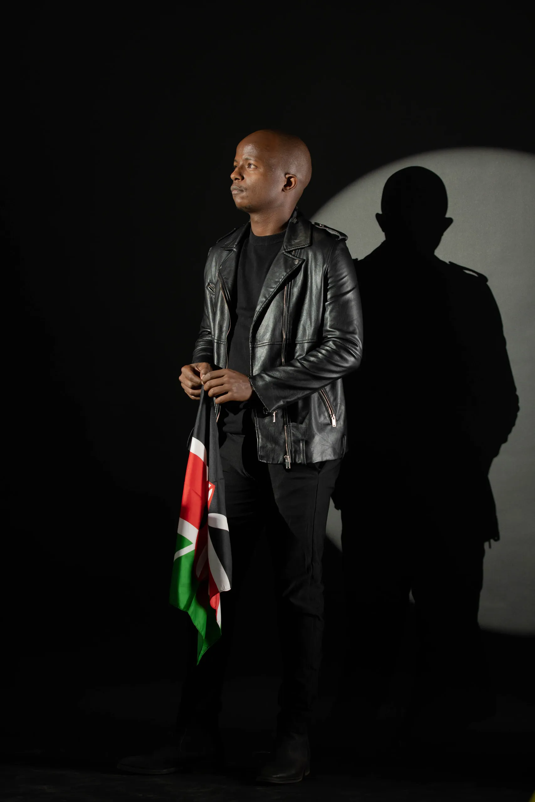 Man in a leather jacket holding a Kenyan flag against a black background, his shadow on a round light
