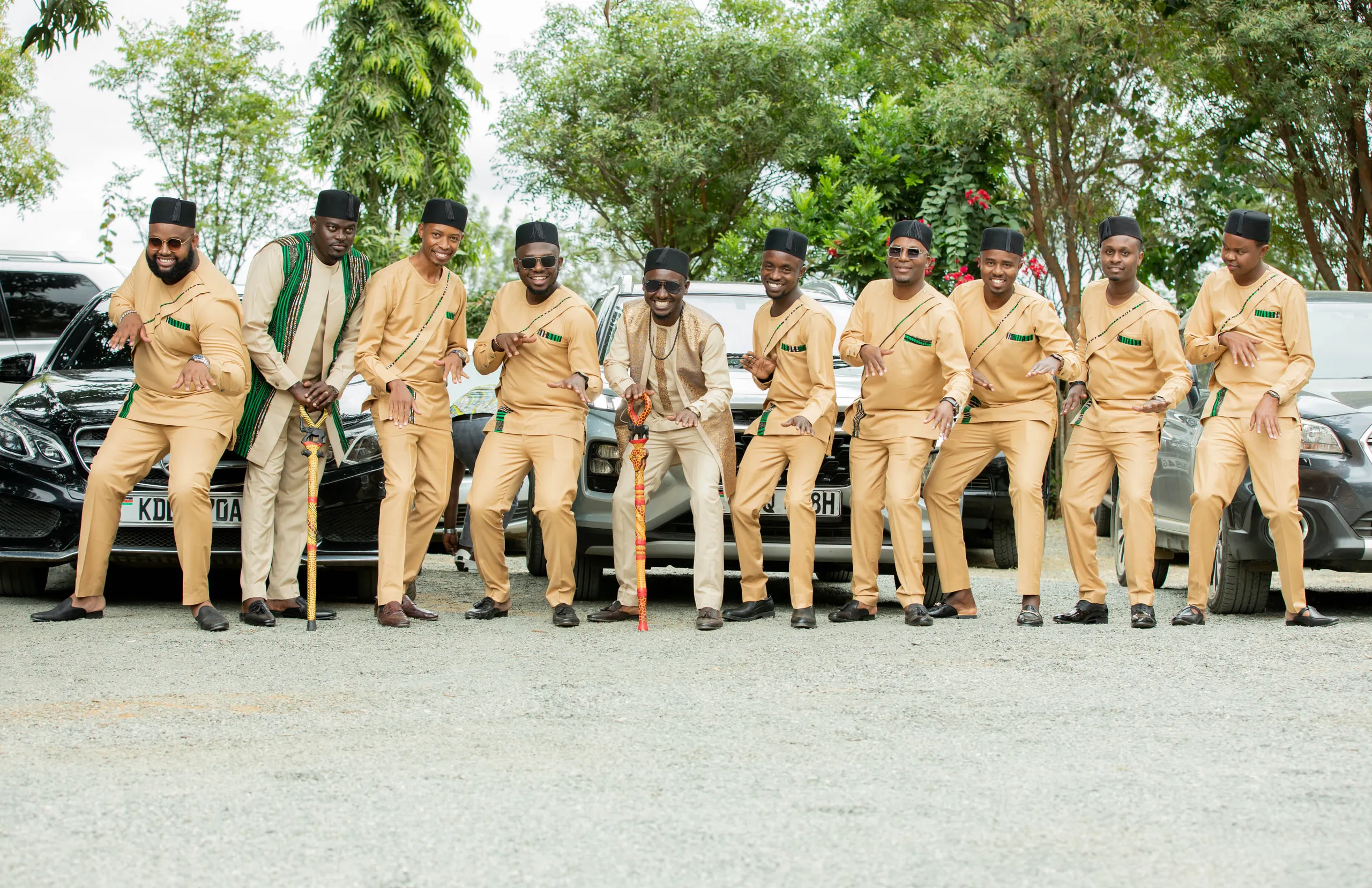 The groom and his party in matching tan outfits dancing in front of the cars