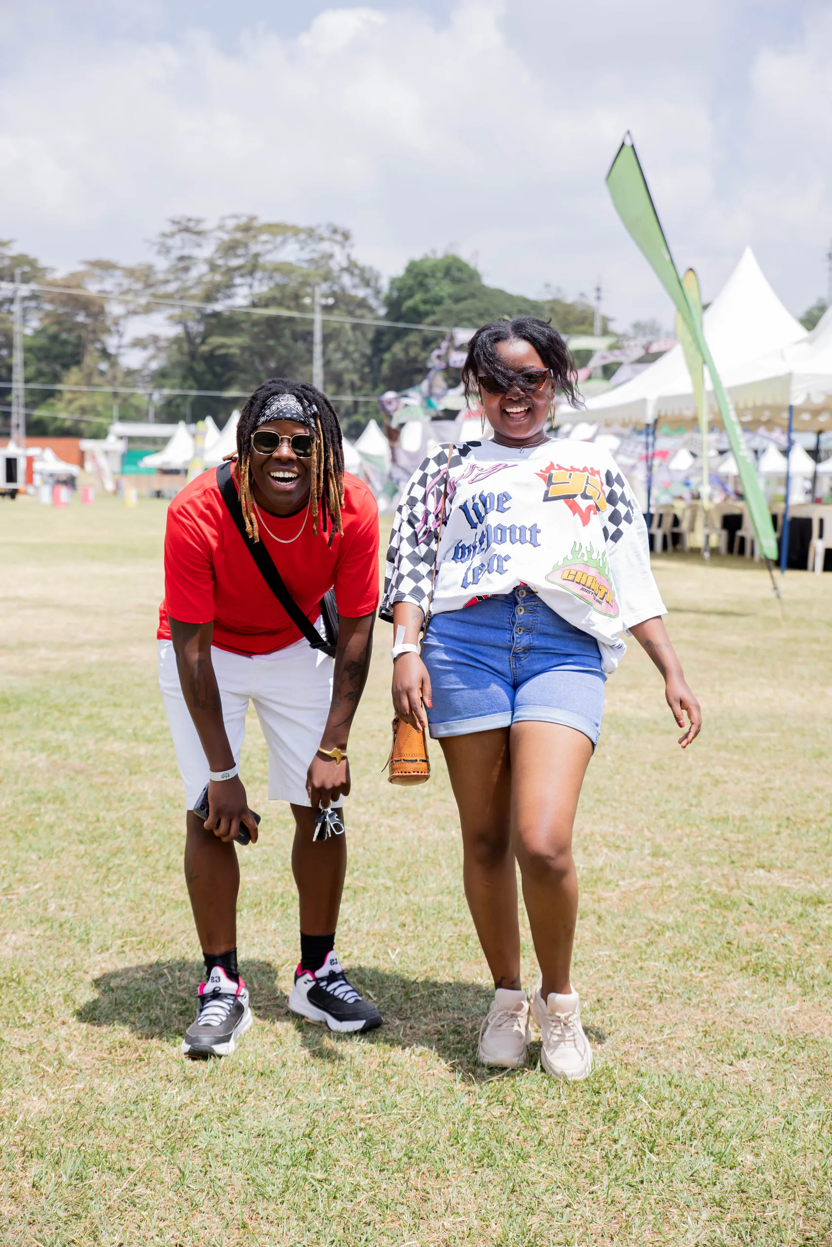 Two friends laughing on the grass, one bent forward, at an outdoor festival