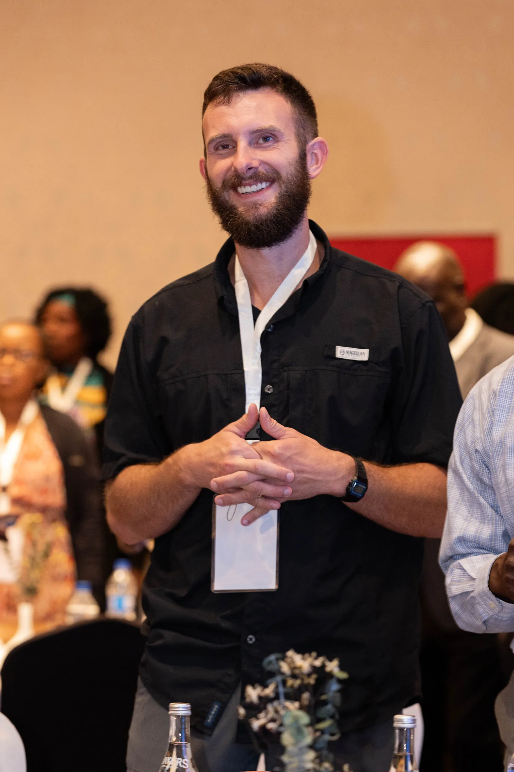 Smiling bearded man with a conference lanyard standing among guests in a ballroom