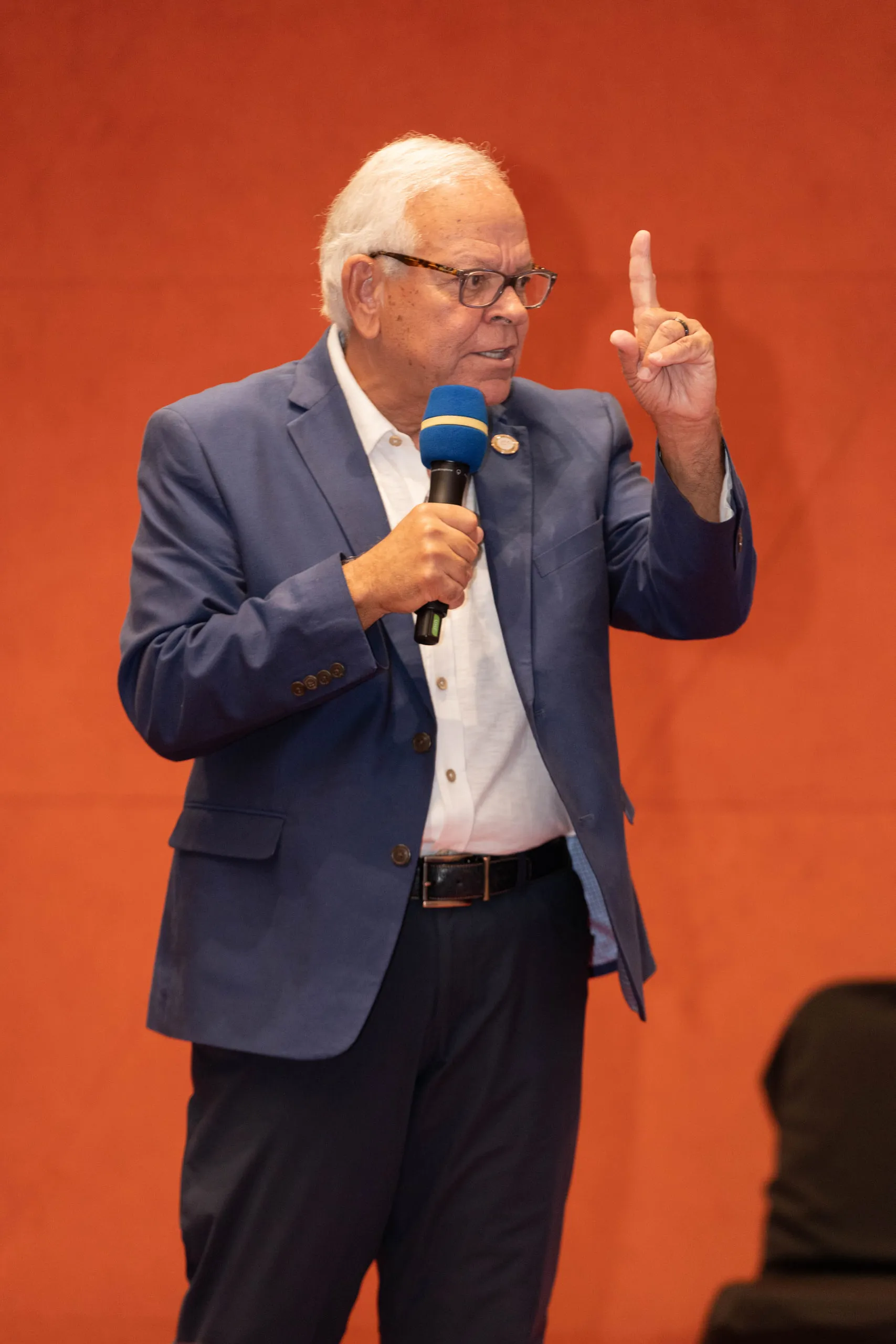 Older man in a navy blazer holding a microphone and raising one finger