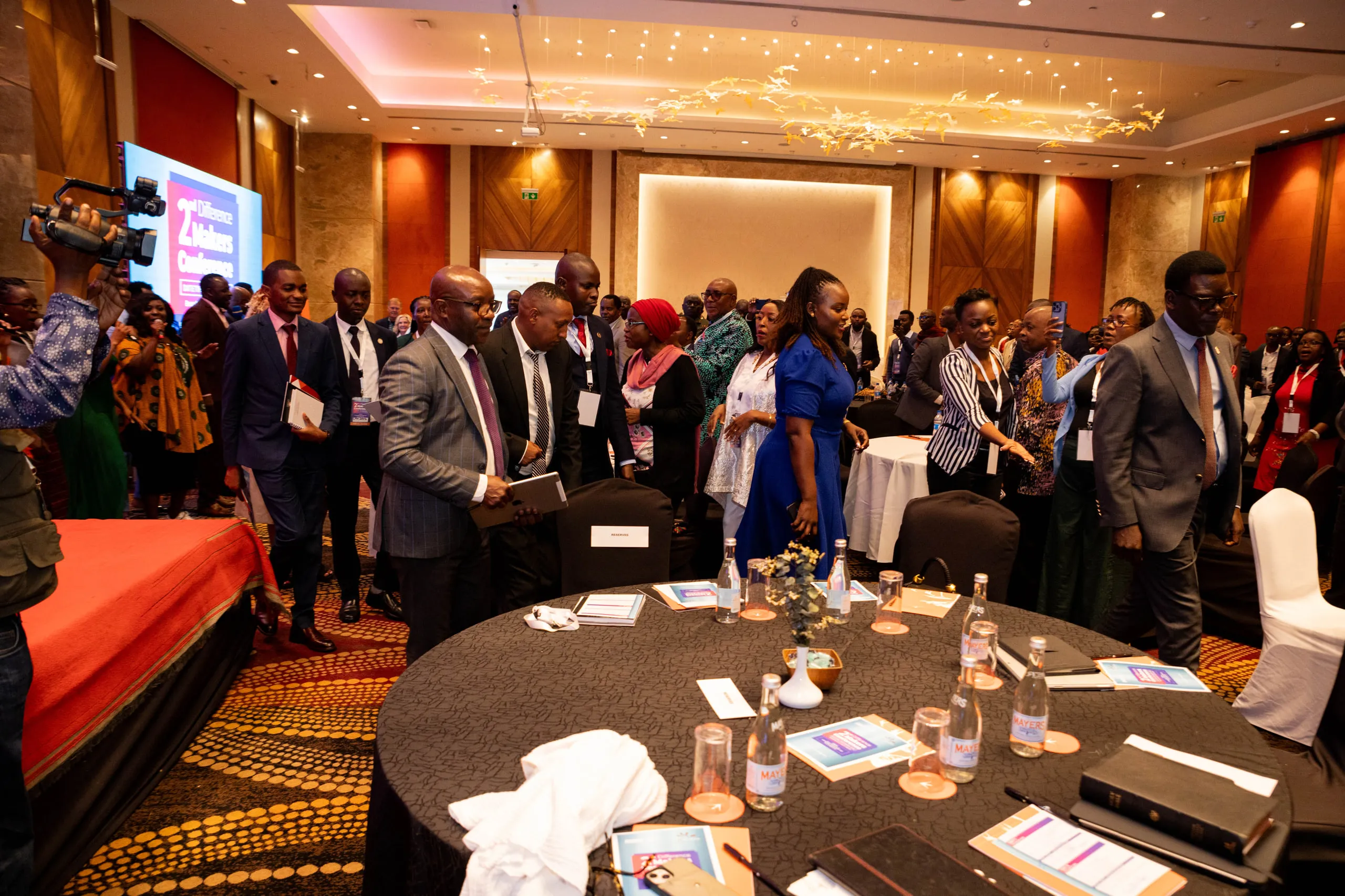 Delegates standing at round tables in a hotel ballroom with a camera operator at left
