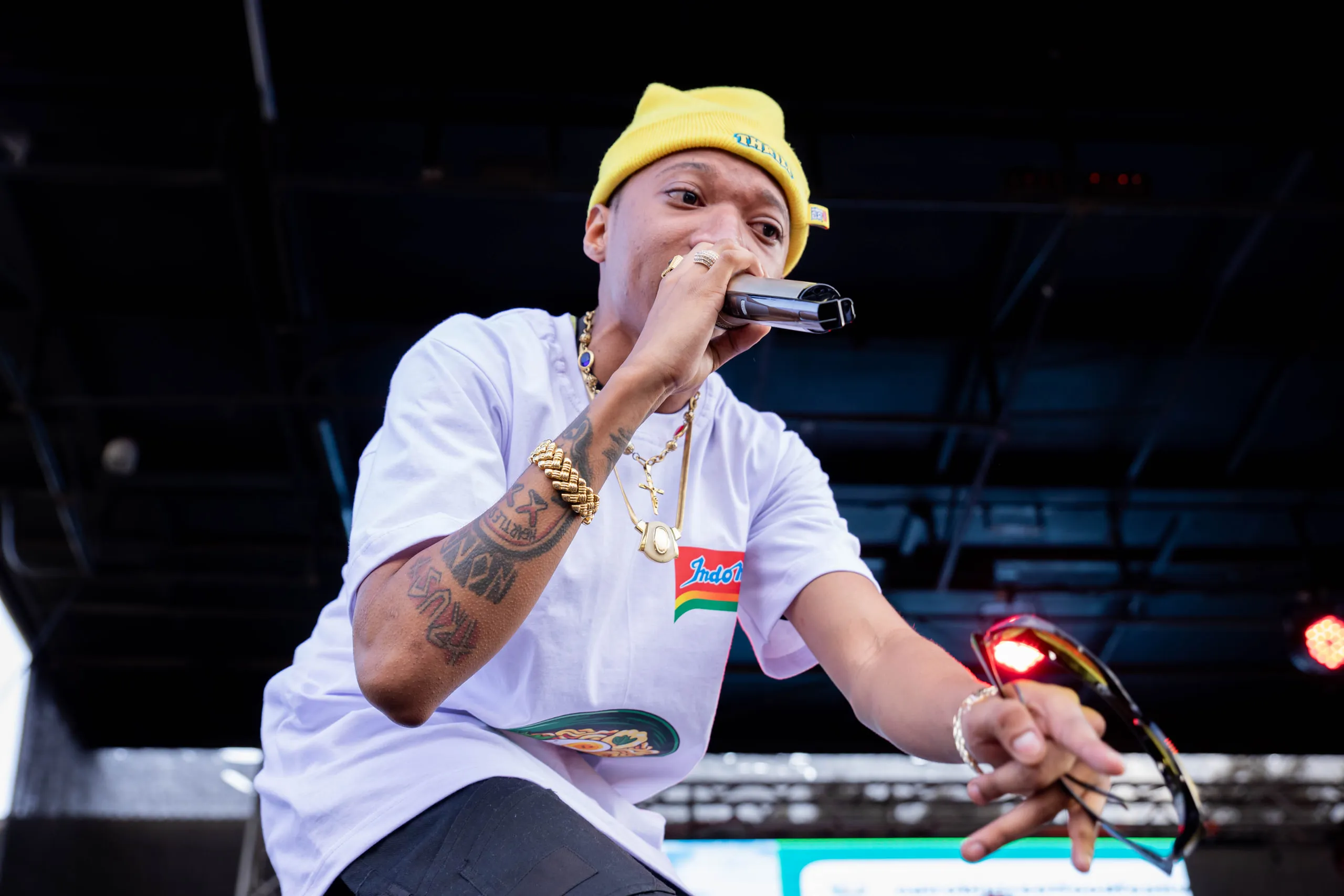 Rapper in a yellow beanie and white tee leaning forward with a microphone on stage