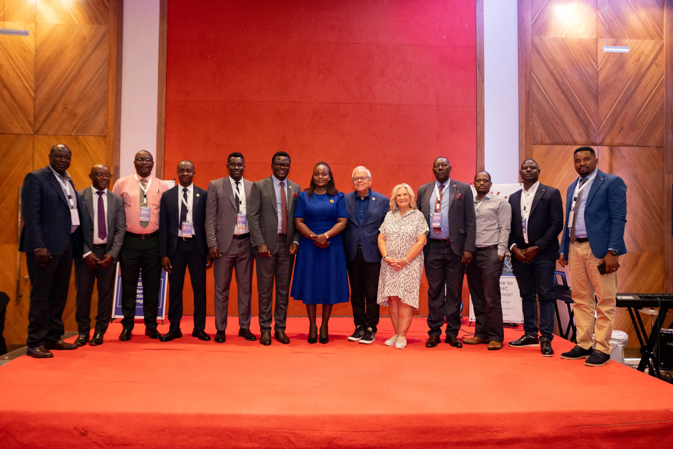 Twelve conference guests lined up on a red stage floor beside a woman in a blue dress