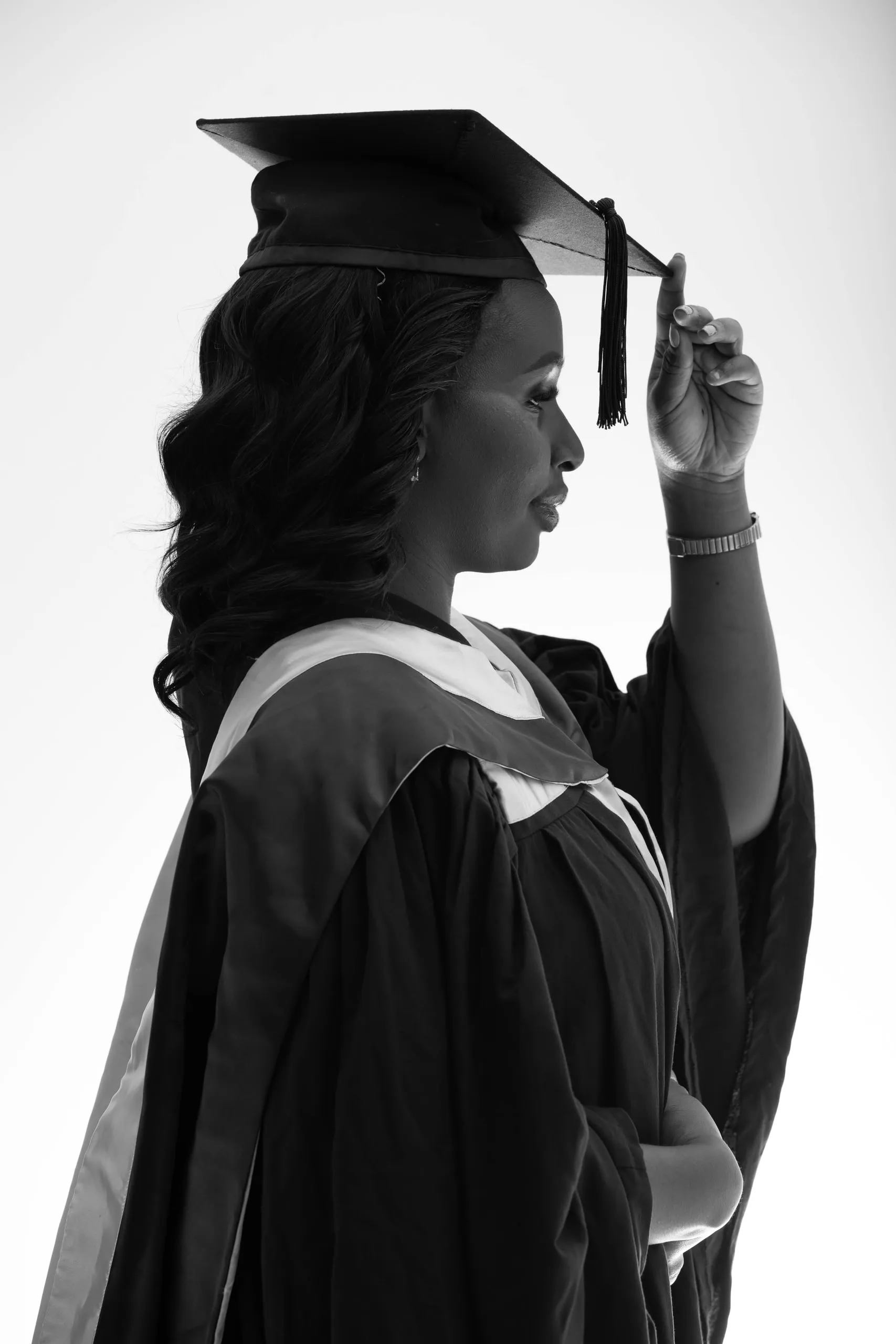 Woman in a graduation gown and cap in profile, touching the brim of the cap, in black and white