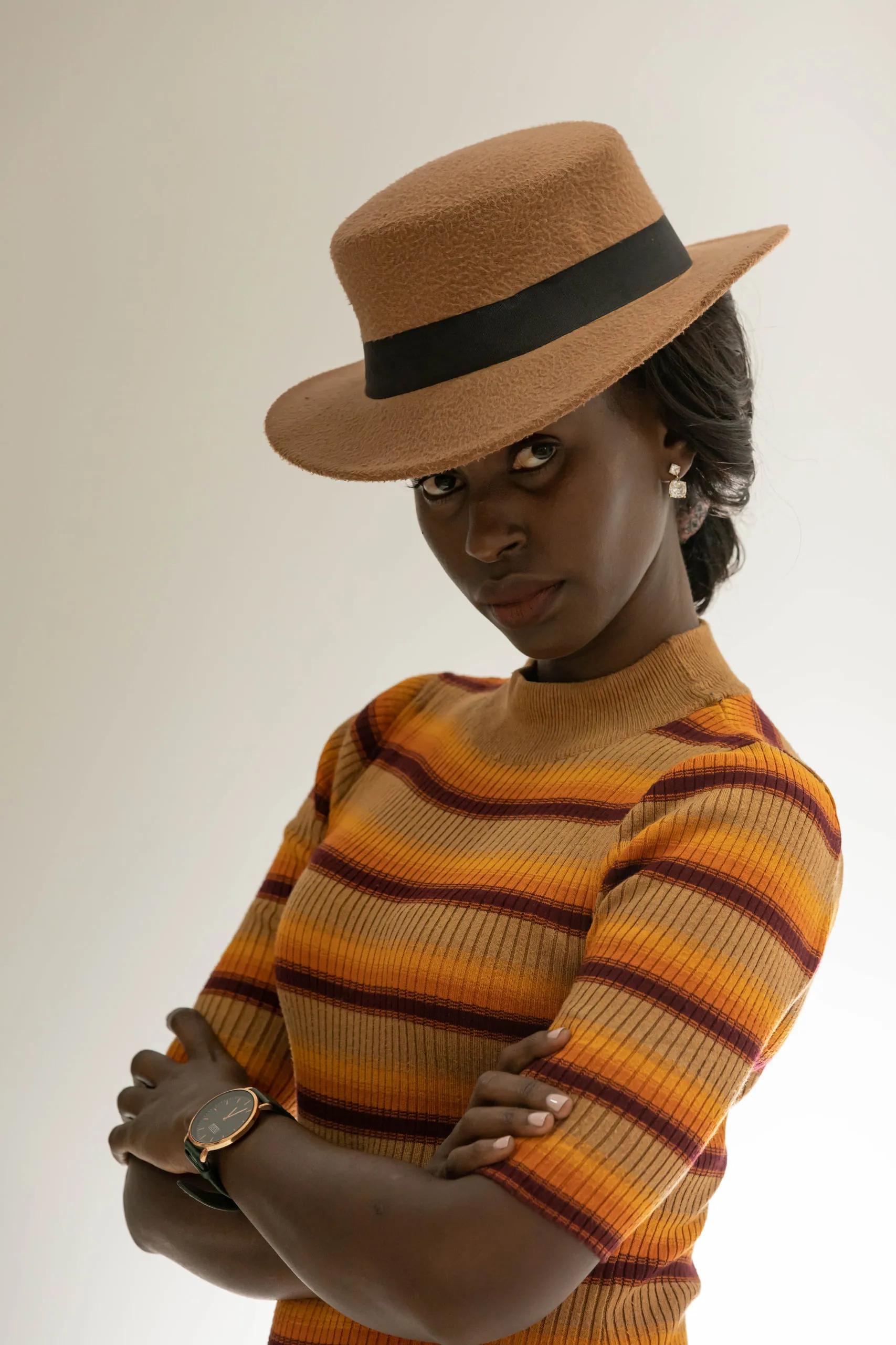 Woman in a brown hat and striped orange knit top with her arms crossed, looking at the camera