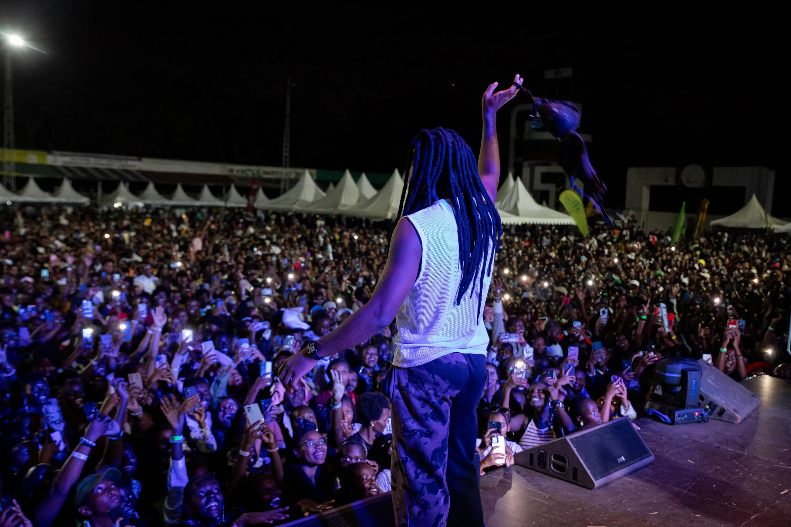 Performer with long braids and a raised arm facing a packed night crowd holding up phone lights