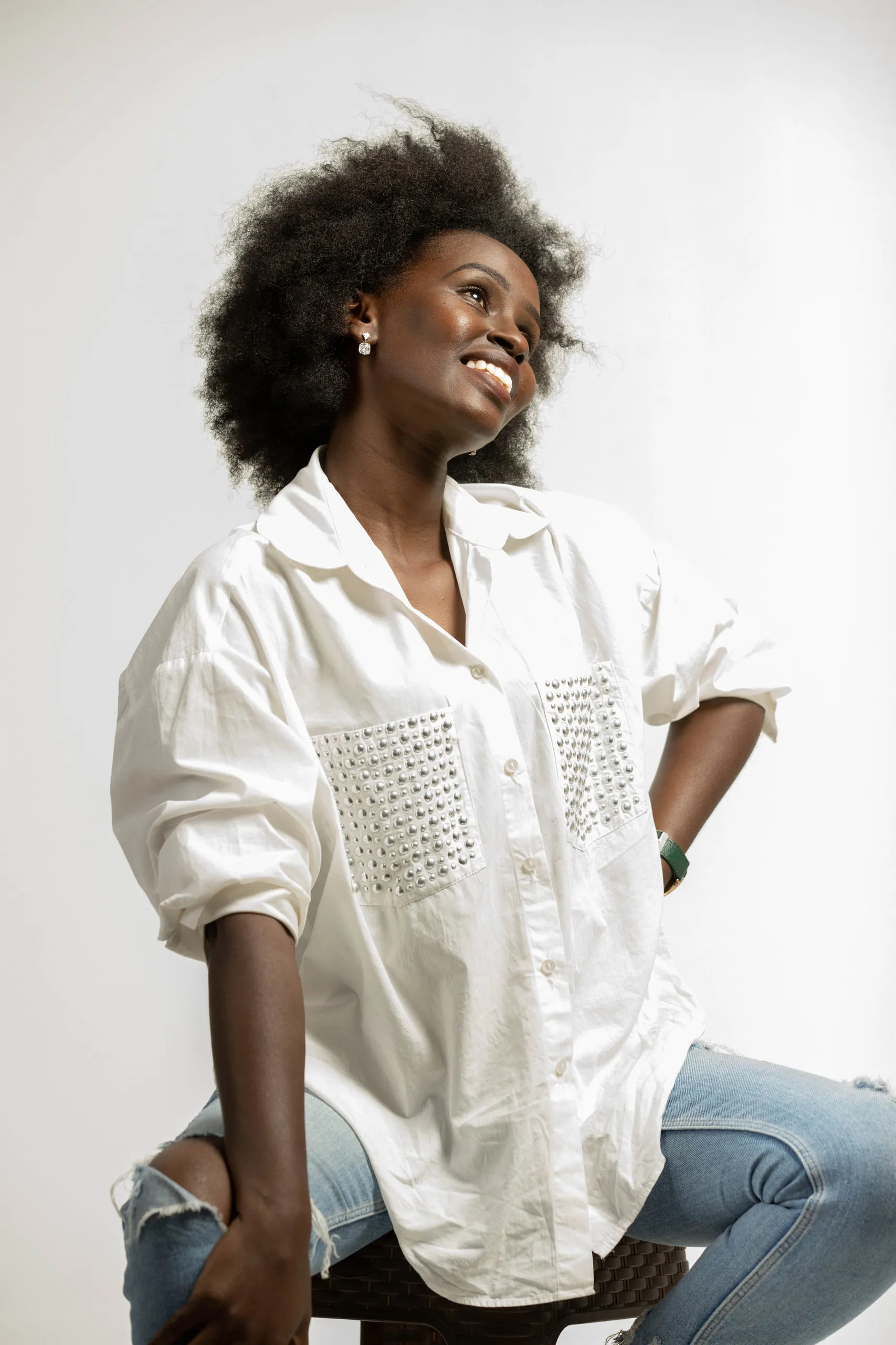 Woman with an afro in an oversized white shirt looking up and smiling, sitting on a stool