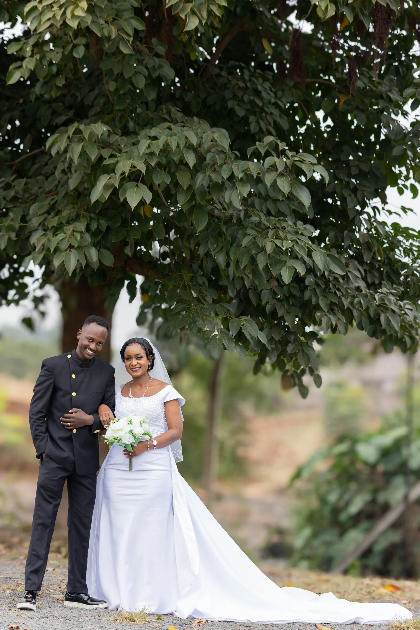 Bride and groom under a tree, her long train across the gravel