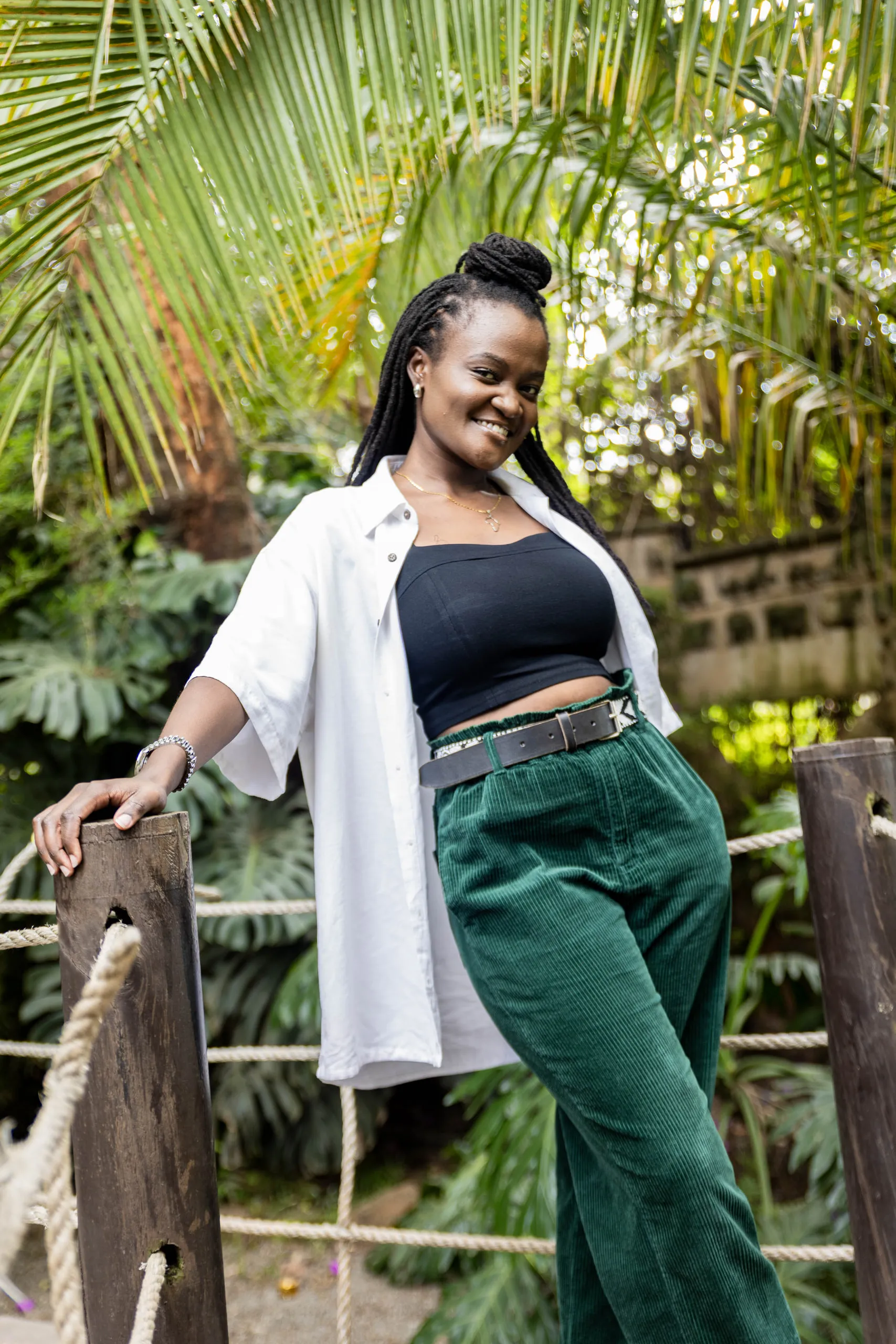 Woman in a white shirt and green corduroy trousers leaning on a rope fence under palm leaves