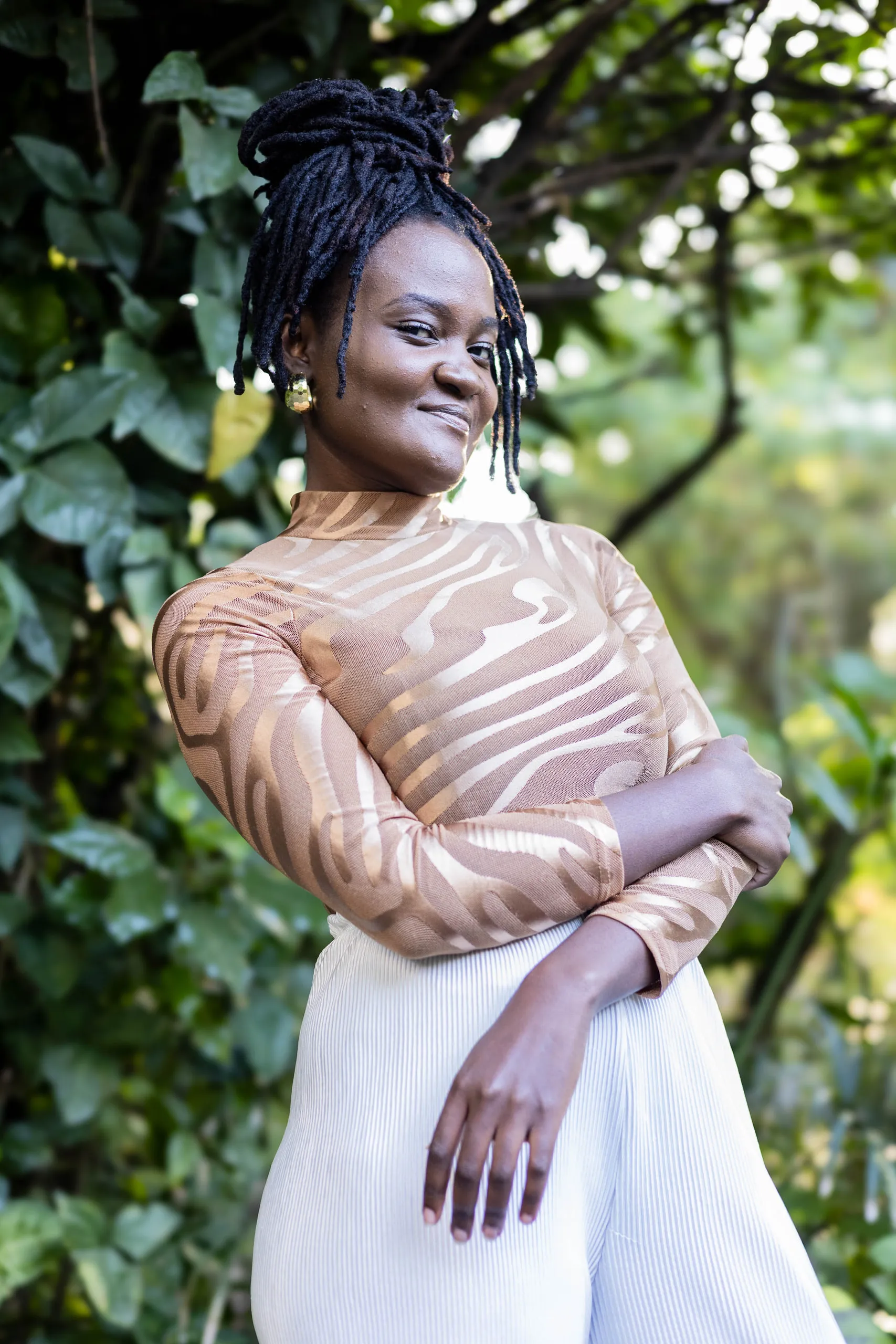 Woman with a high bun of locs in a gold-patterned top, arms crossed, against green foliage