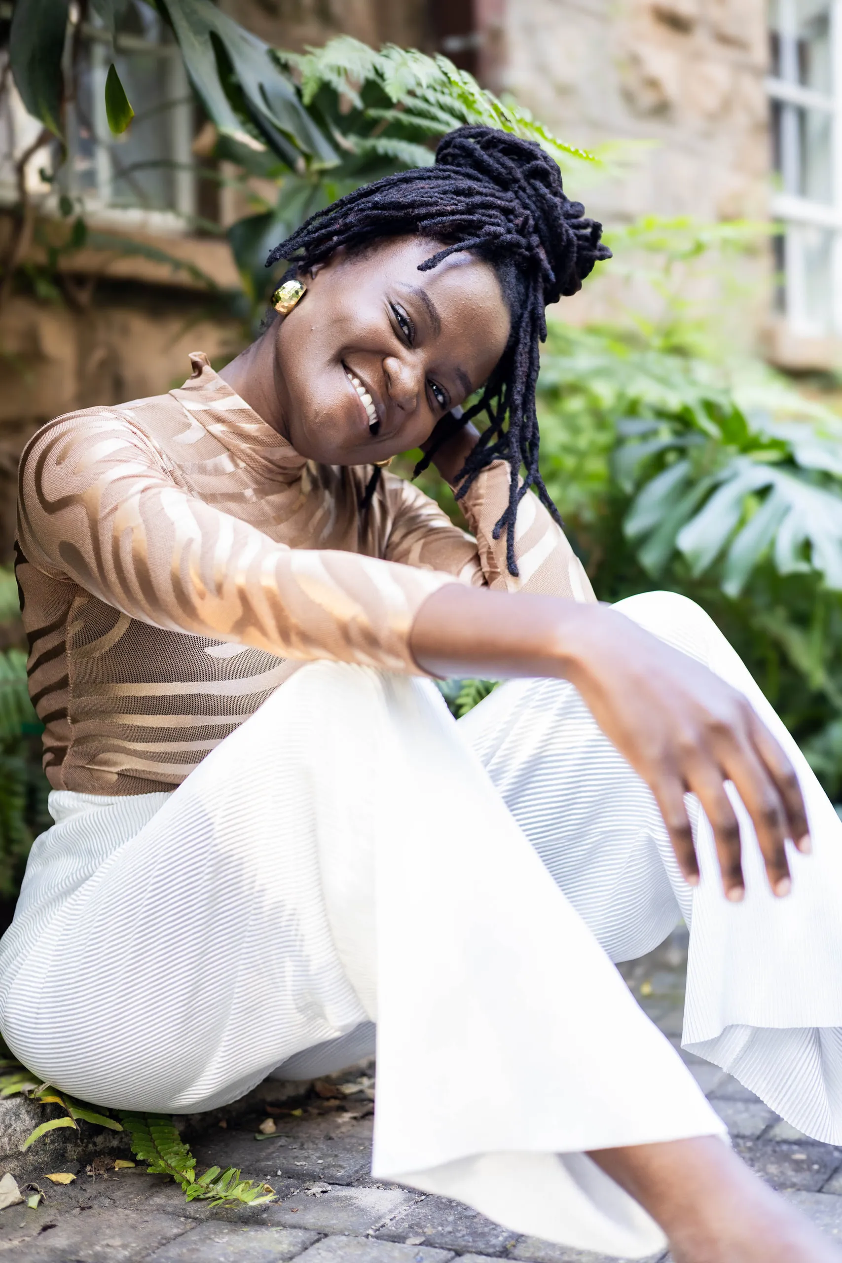 Woman with locs in a patterned top and white trousers, tilting her head and smiling on a stone path