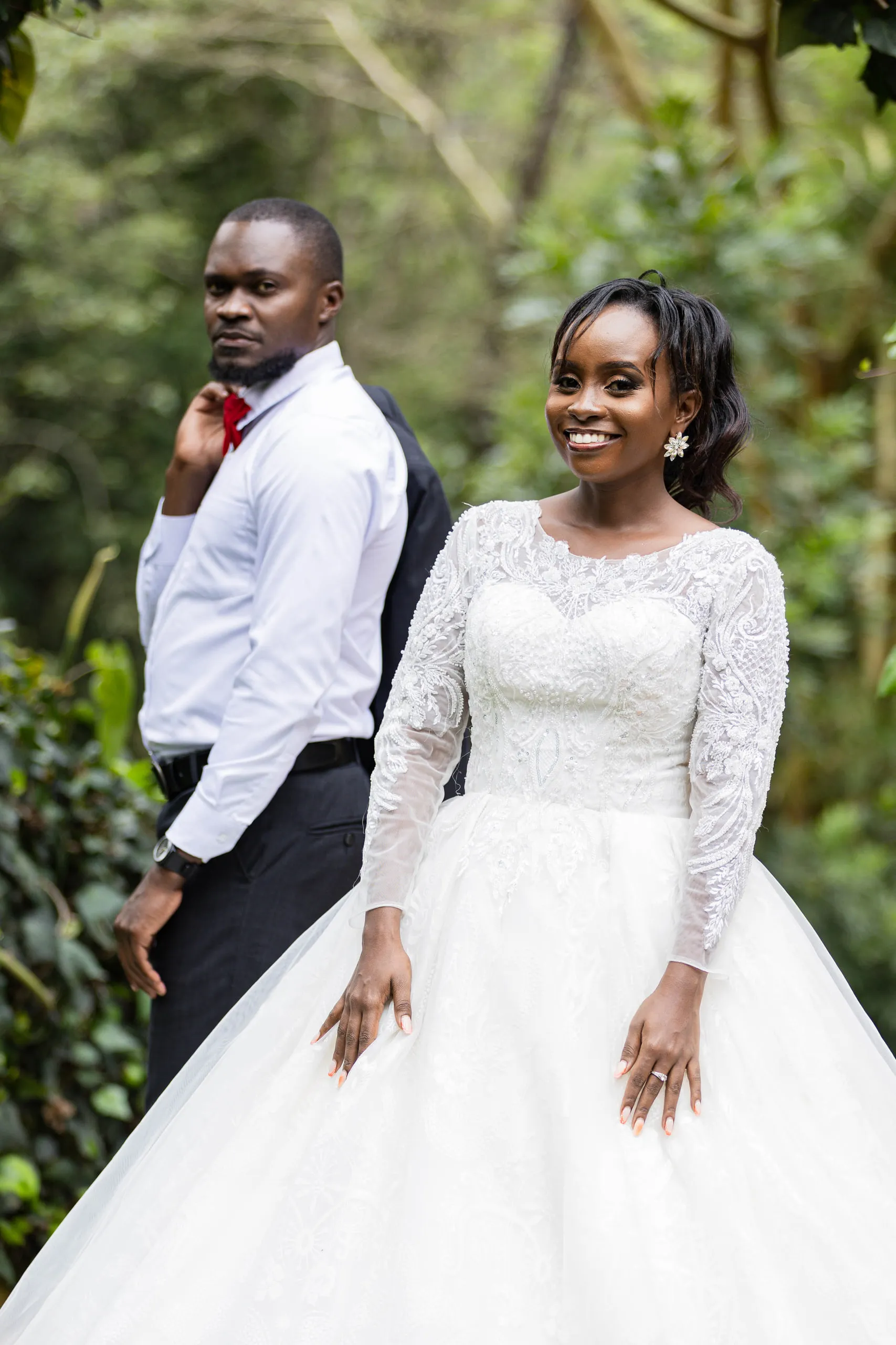 Smiling bride in a long-sleeved lace gown, the groom behind her