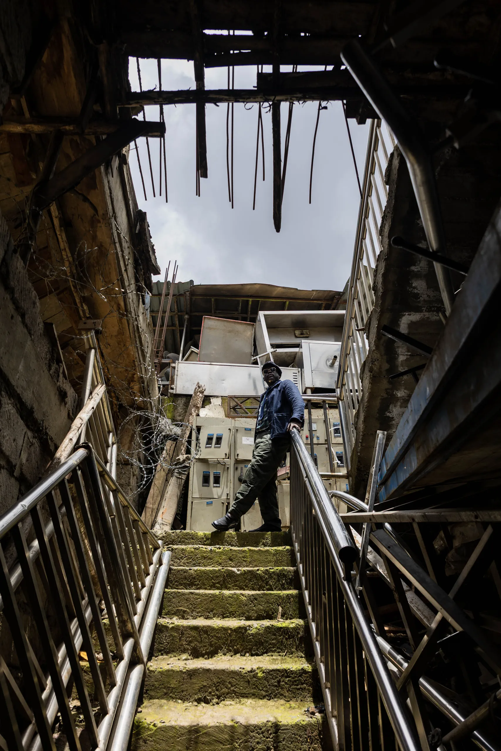 Man in a blue jacket standing on a mossy staircase in a derelict building, seen from below