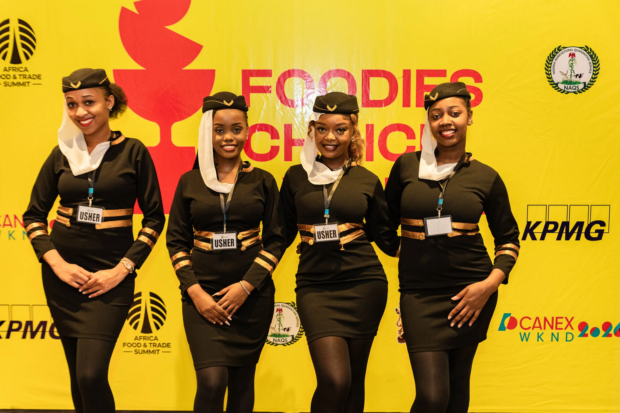 Four ushers in black uniforms and caps smiling in front of a yellow event backdrop