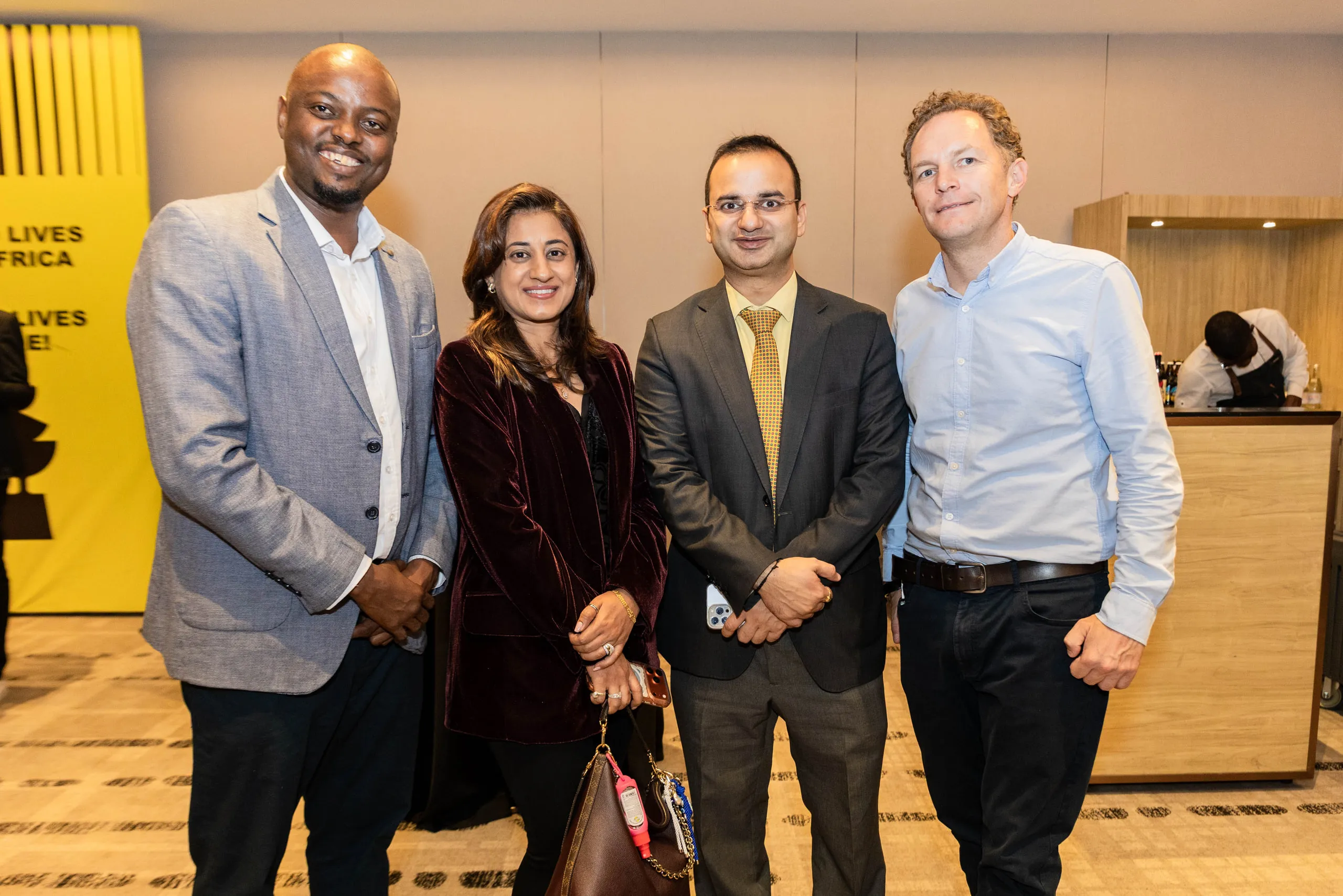 Four guests posing together beside a yellow event banner in a hotel lounge