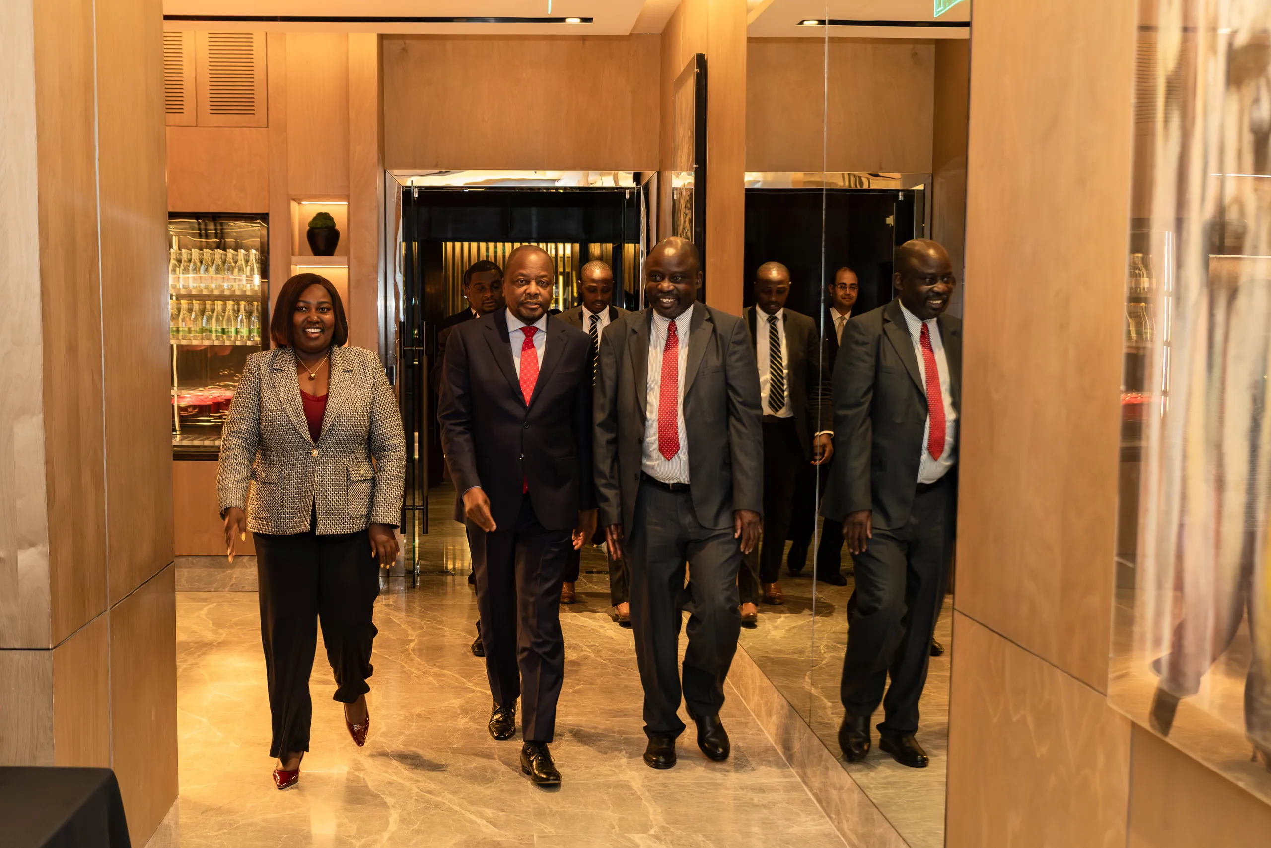 Five people in suits walking toward the camera through a hotel passage with mirrored walls