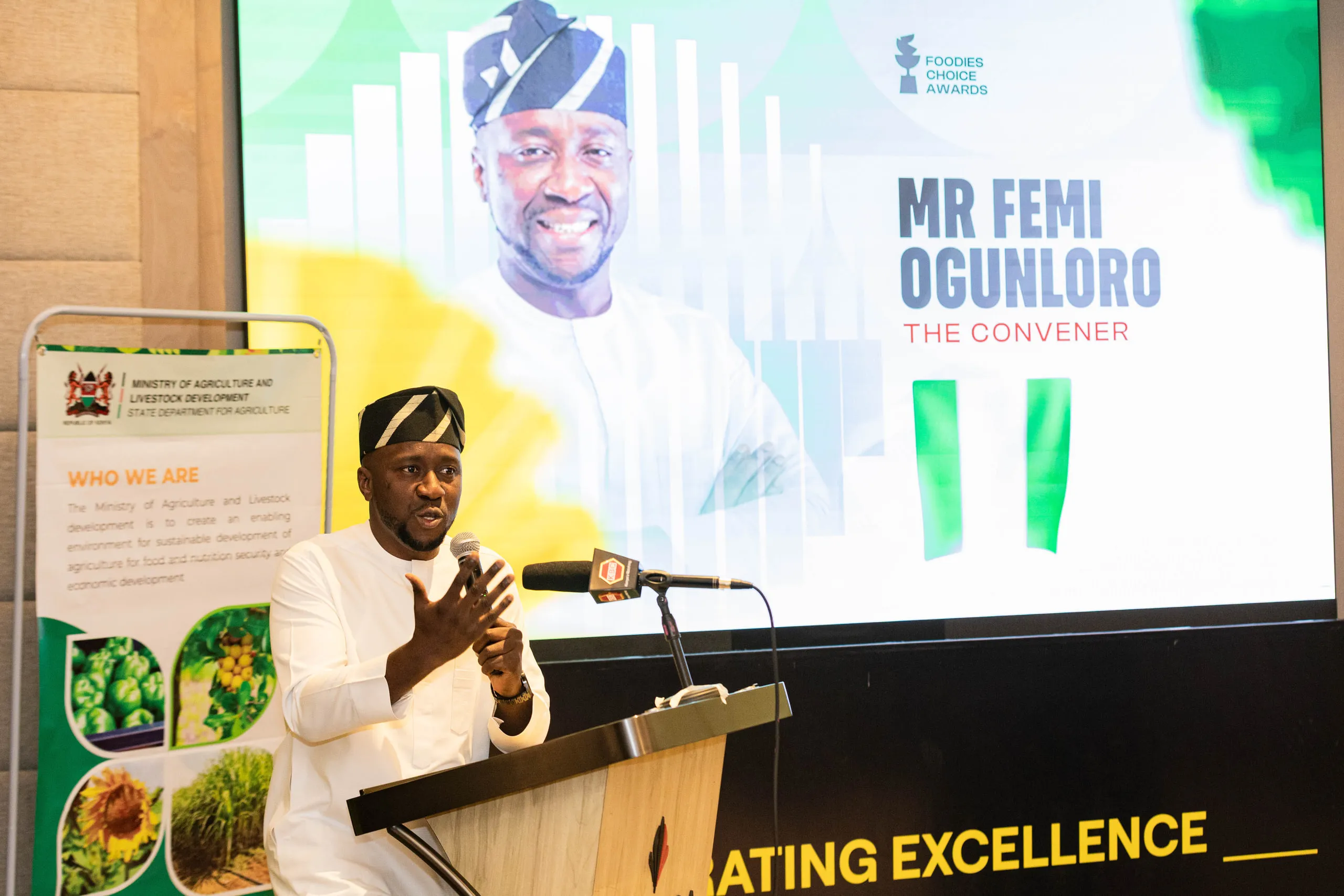 Man in white traditional dress and cap speaking at a lectern in front of a large screen