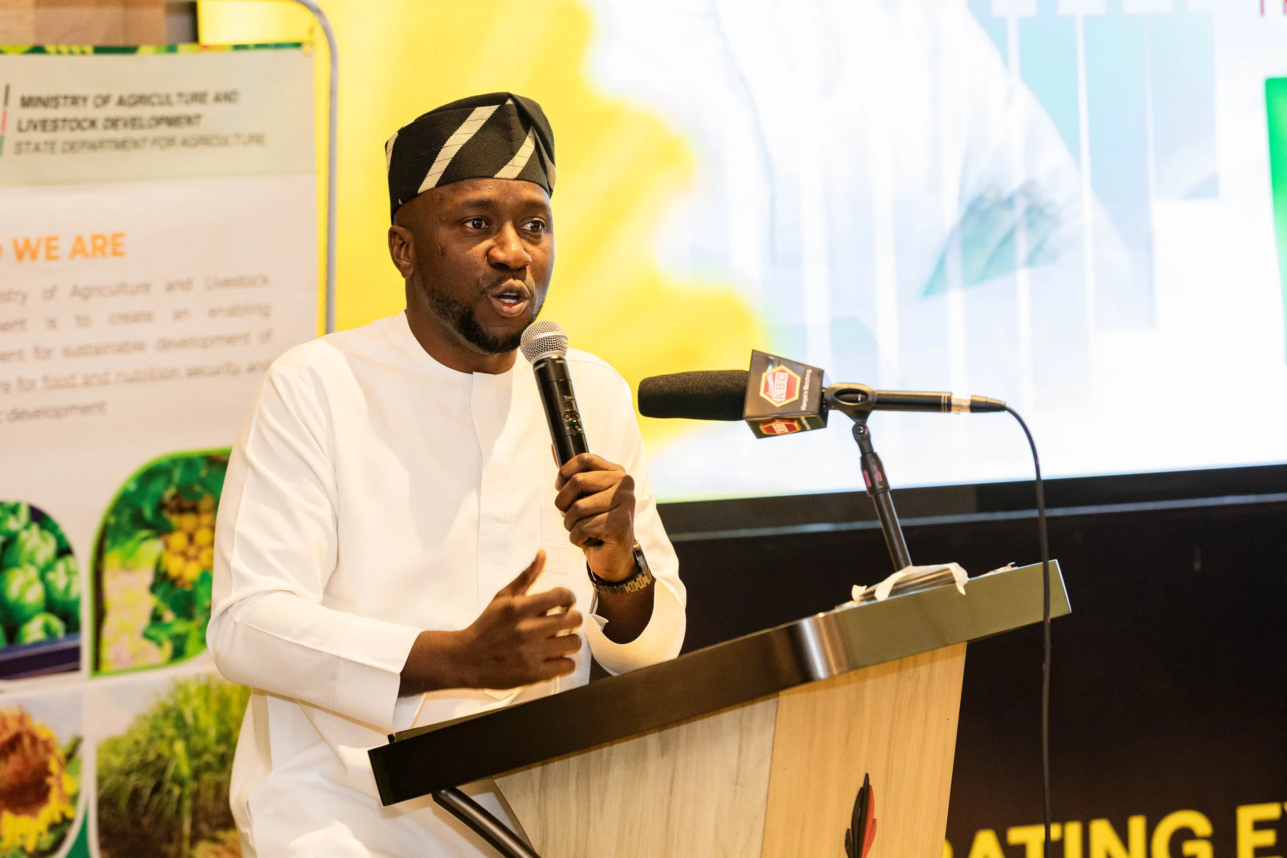 Man in a white outfit and patterned cap speaking into a handheld microphone